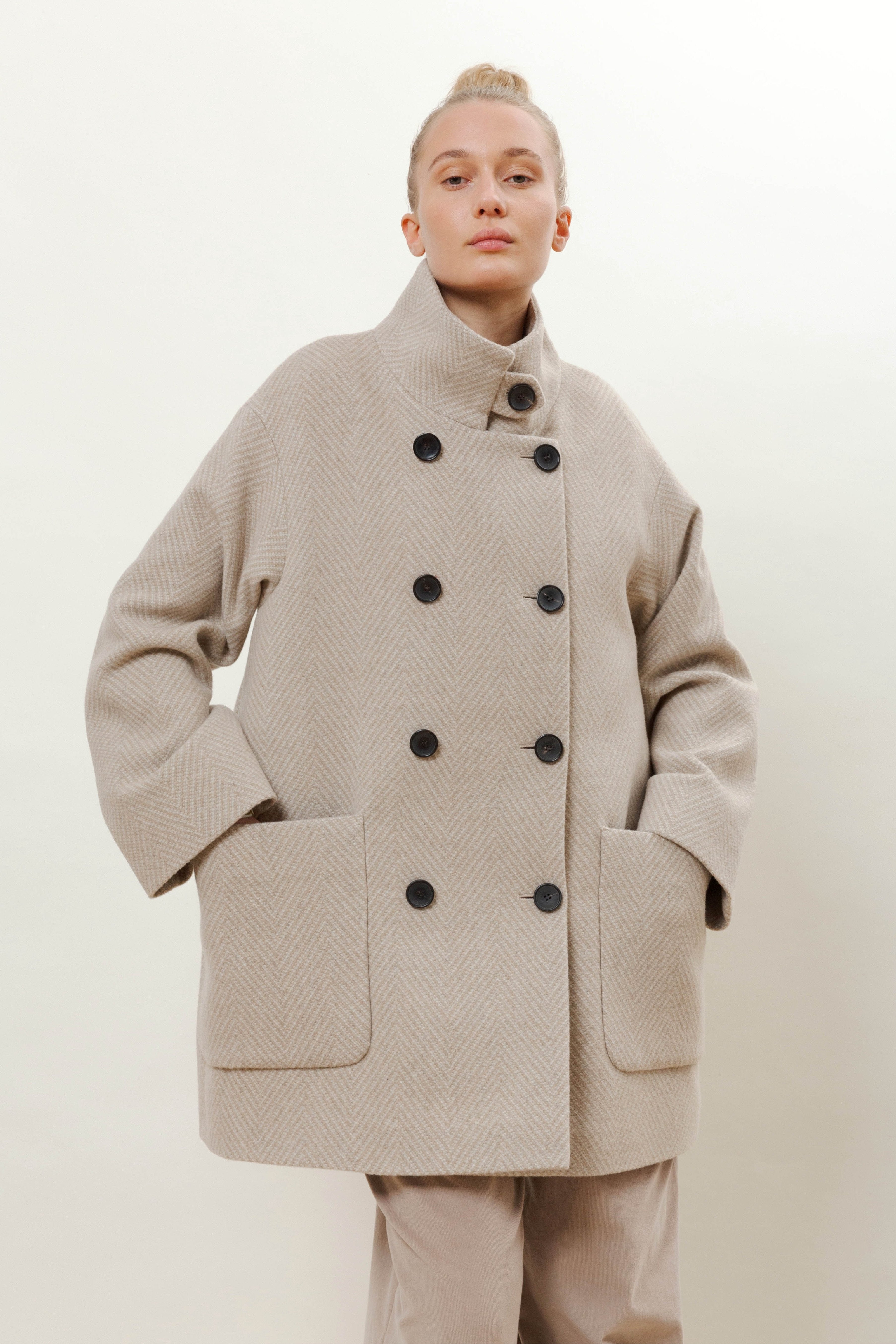 Woman in beige herringbone patterned wool jacket with black buttons, pockets and stand collar