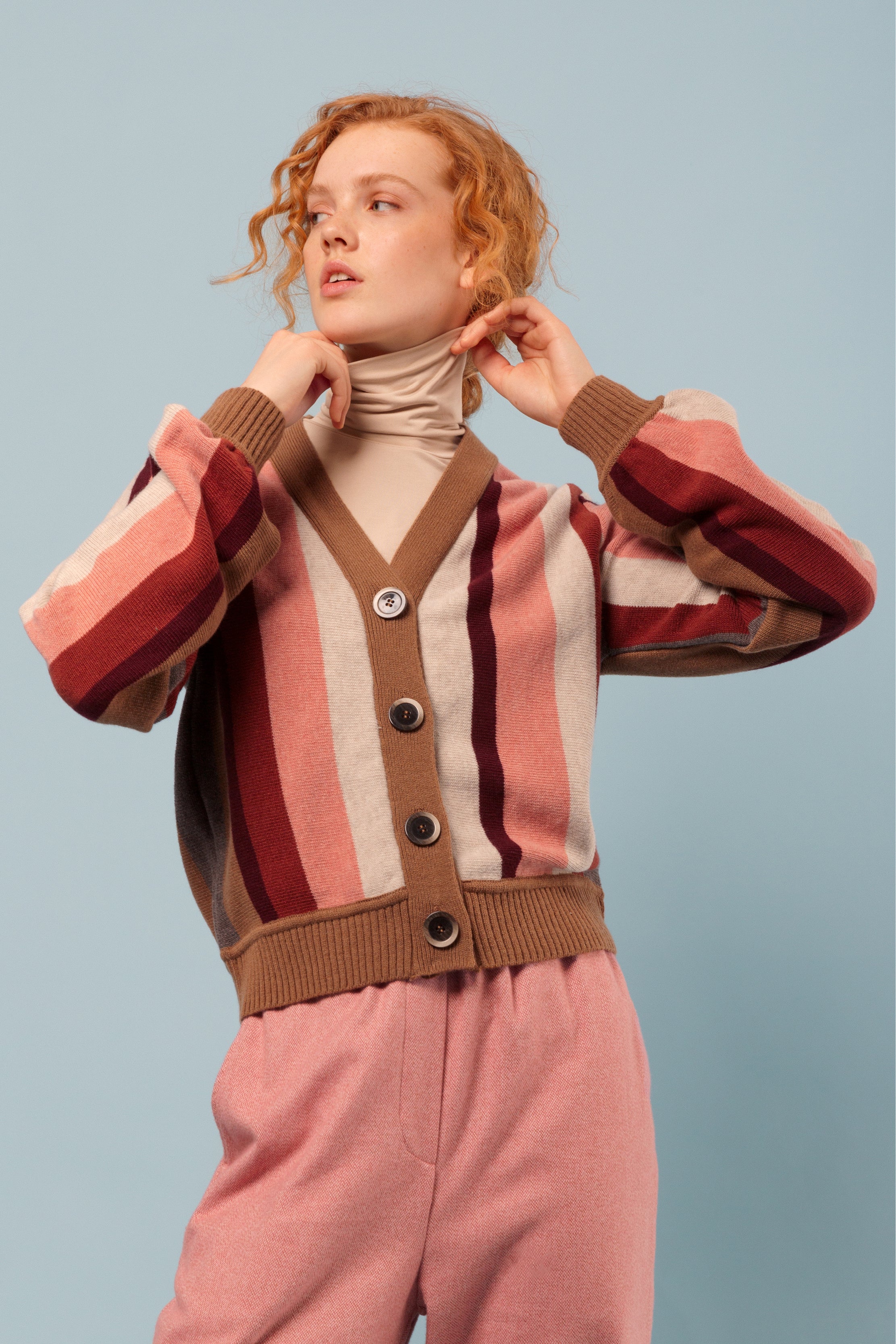 Woman in striped cardigan in brown, beige and pink and with buttons, roll neck shirt and pink trousers