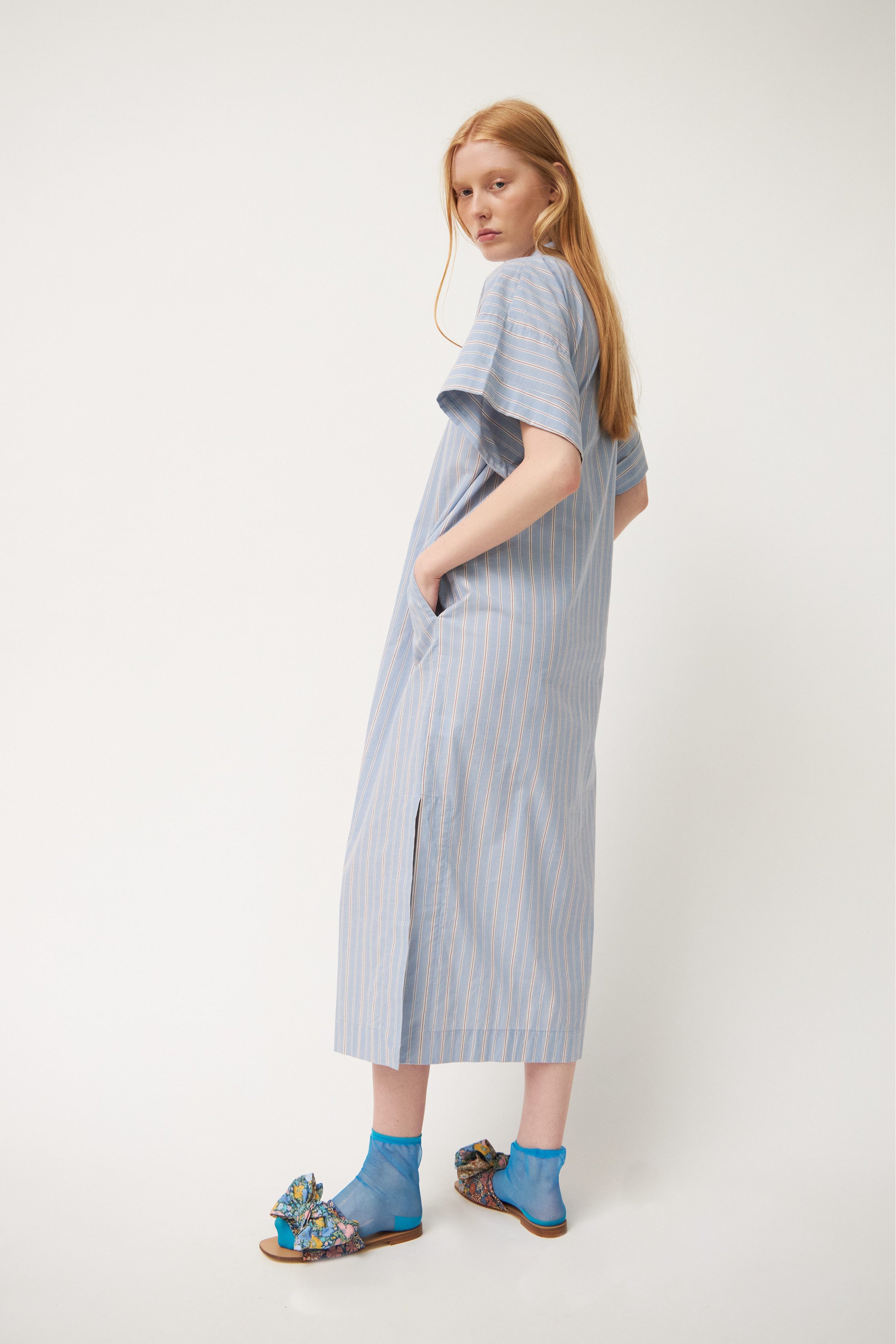 Model wearing blue white and red striped kaftan dress with short sleeves and buttons, blue socks and sandals with flower patterned bows