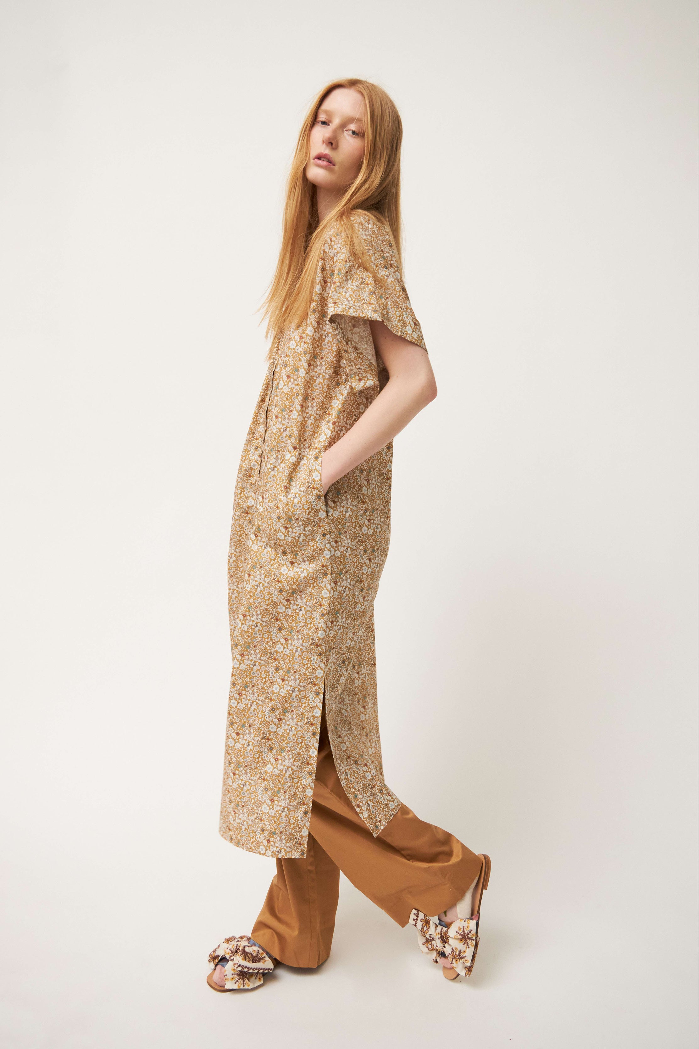 Model wearing white and brown flower patterned kaftan dress with short sleeves and buttons, brown pants and sandals with bows