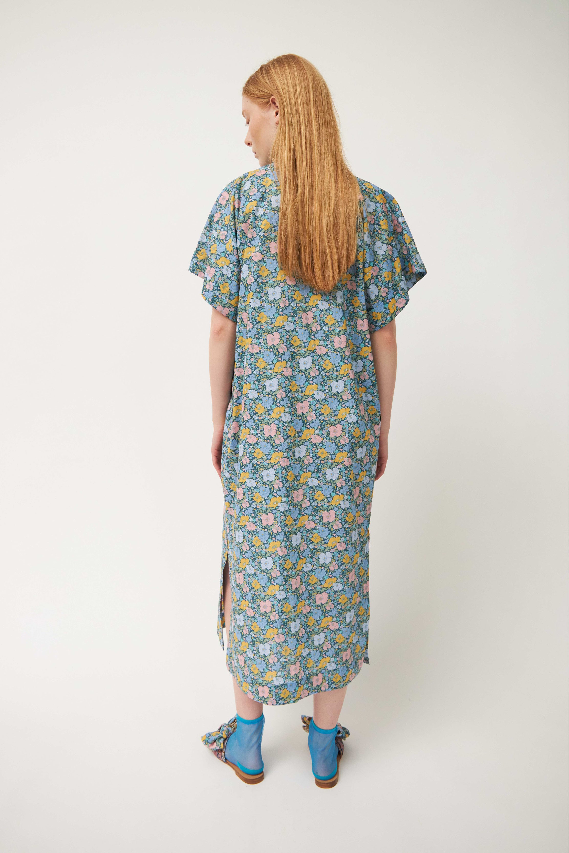 Woman in green multi colored flower patterned kaftan dress with short sleeves and buttons, blue socks and sandals with bows