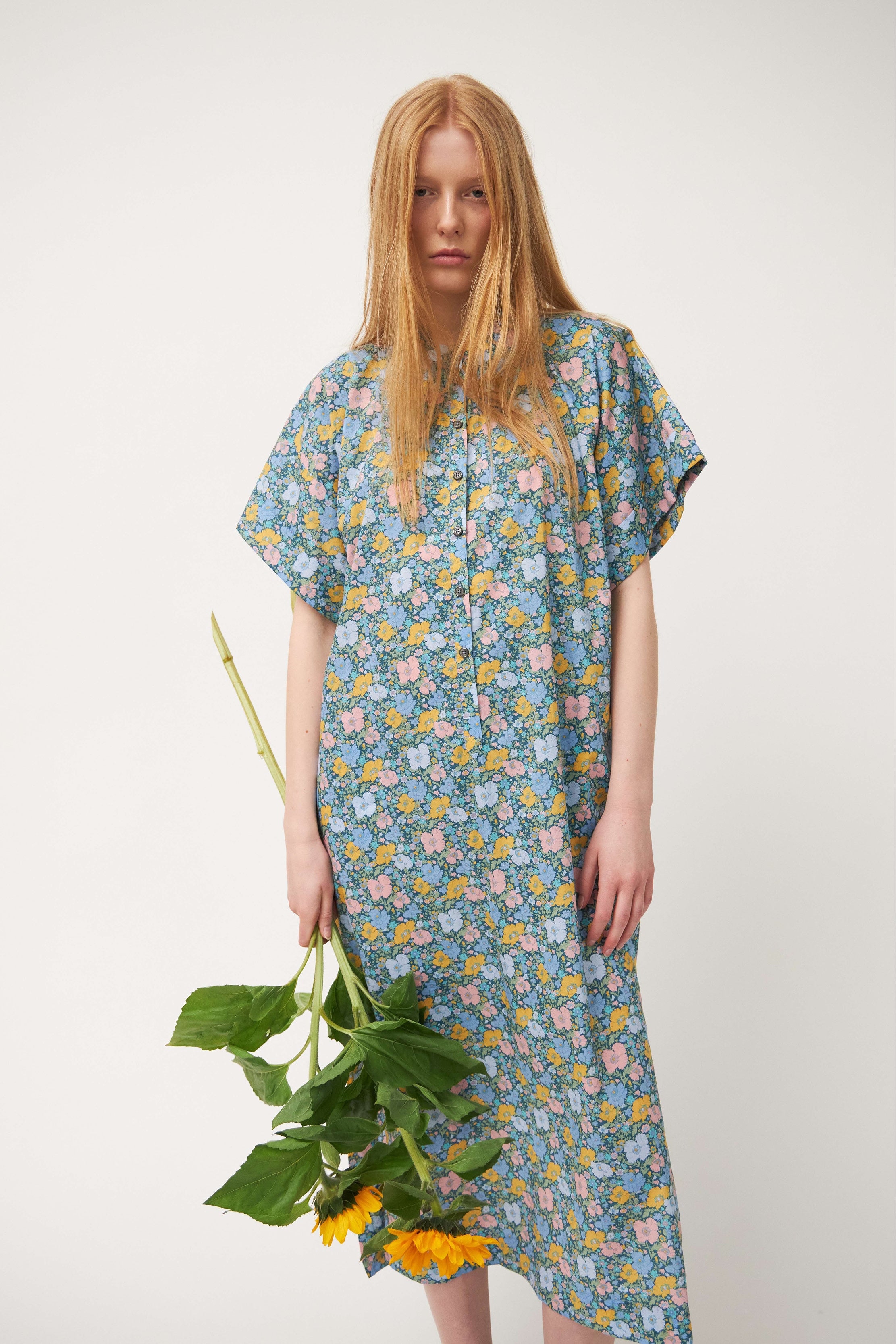 Woman in green multi colored flower patterned kaftan dress with short sleeves and buttons holding sunflowers