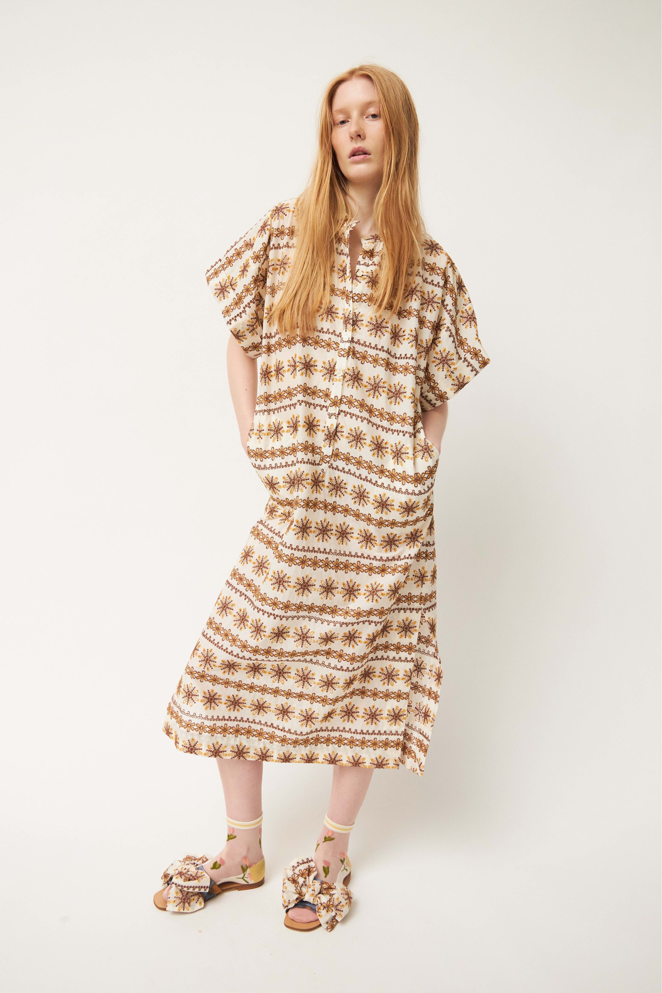 Woman in laced white and brown patterned kaftan dress with short sleeves and buttons, flower patterned socks and sandals with bows