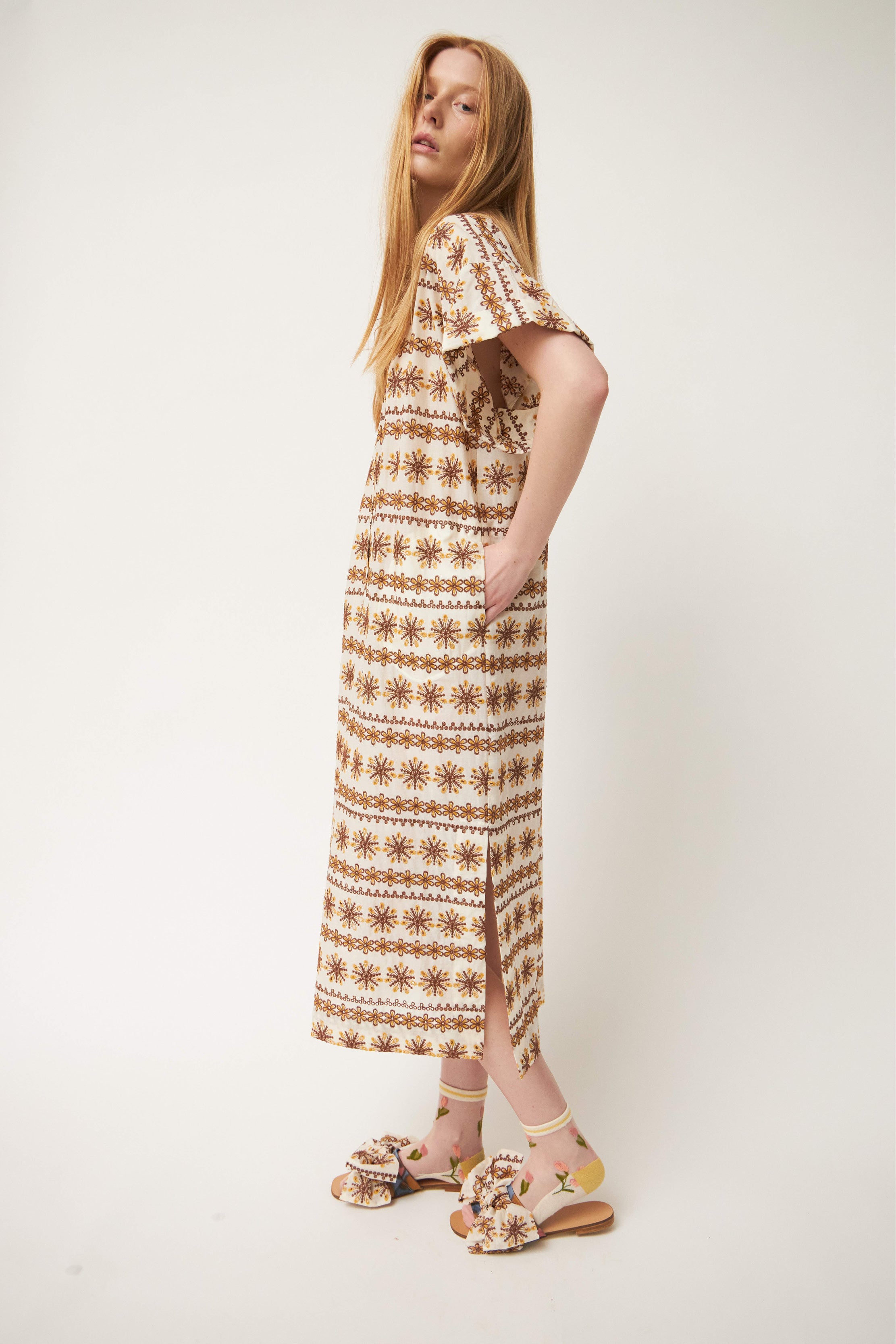 Woman in laced white and brown patterned kaftan dress with short sleeves and buttons, flower patterned socks and sandals with bows