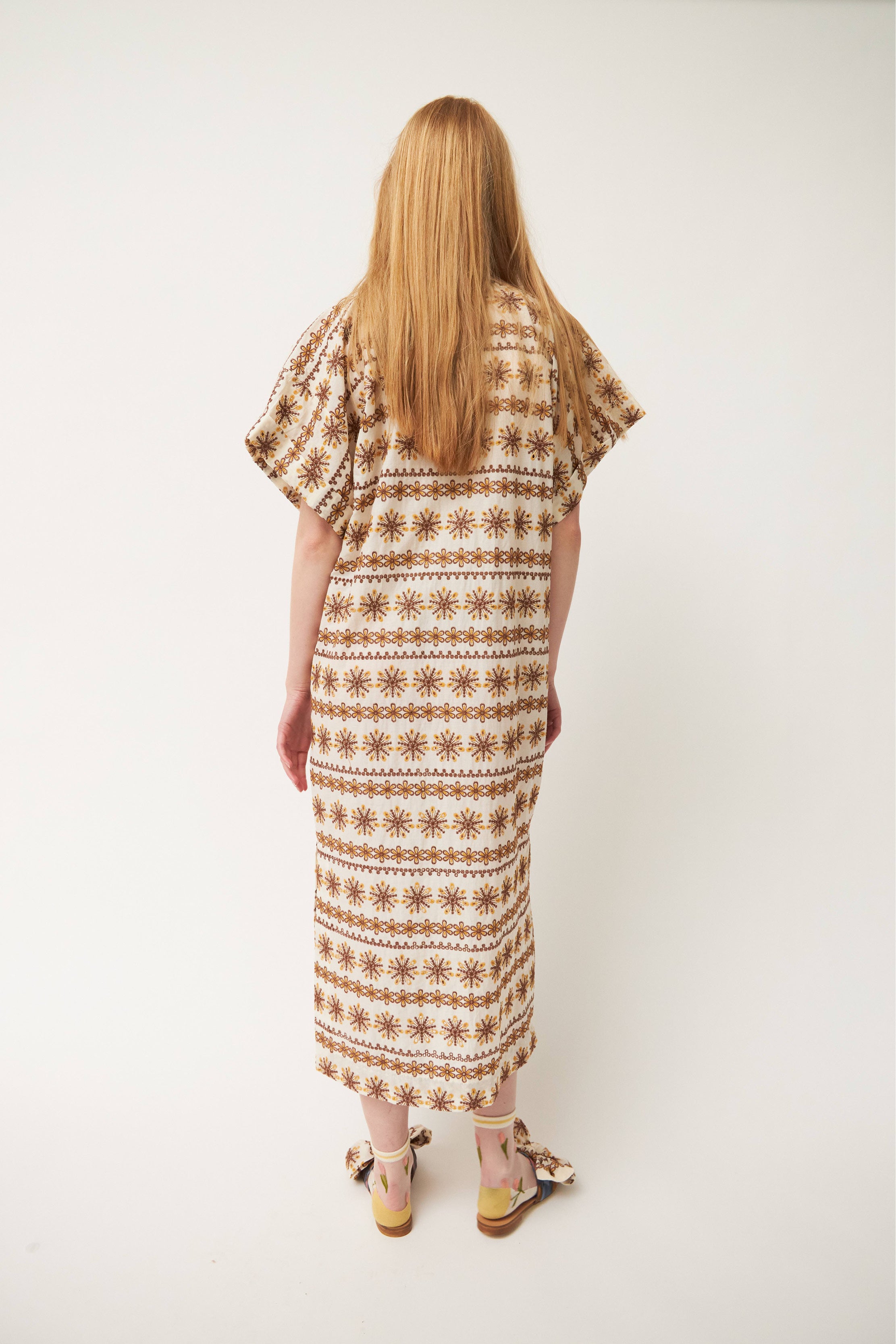 Woman in laced white and brown patterned kaftan dress with short sleeves and buttons, flower patterned socks and sandals with bows