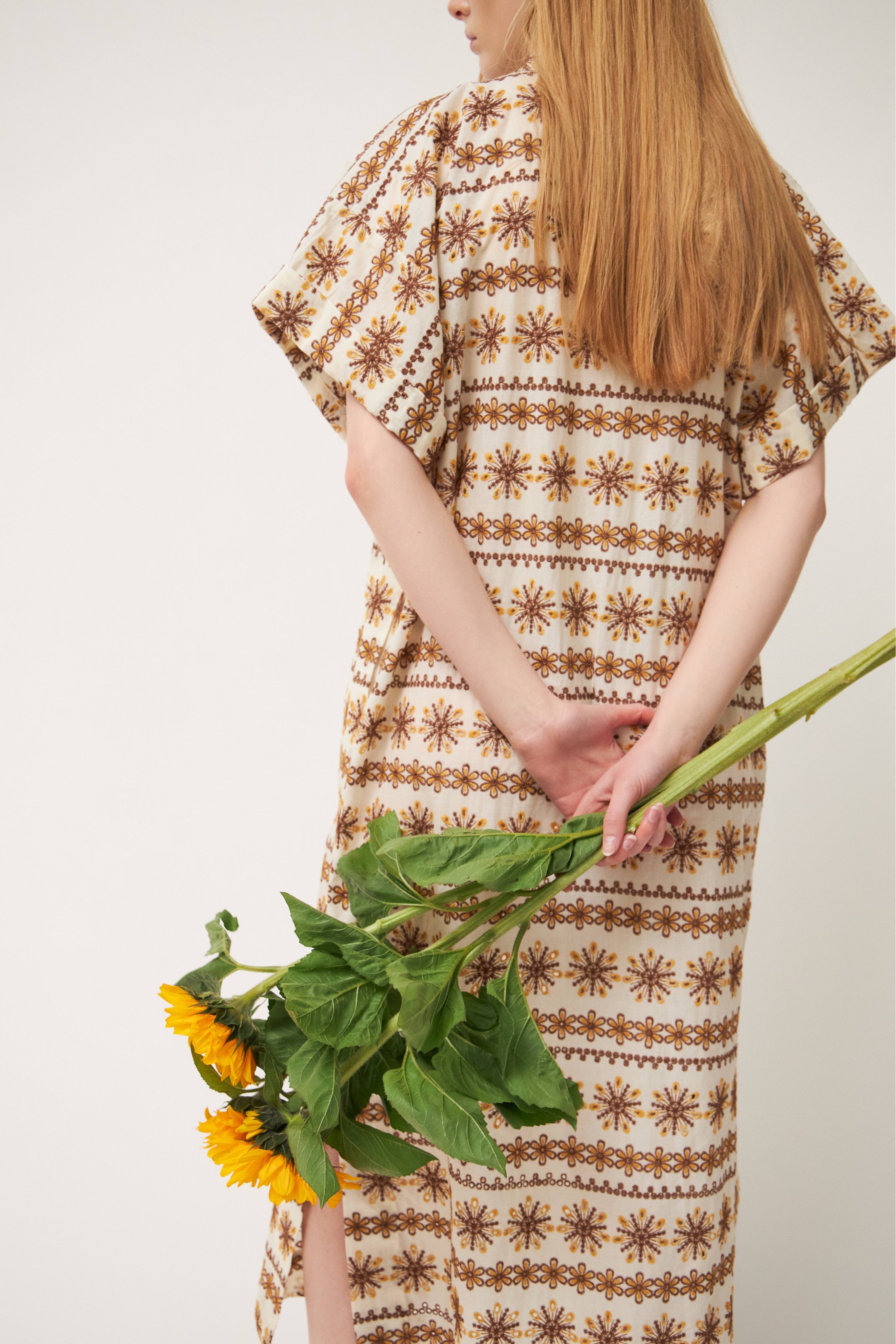 Woman in laced white and brown patterned kaftan dress with short sleeves and buttons holding sunflowers behind back