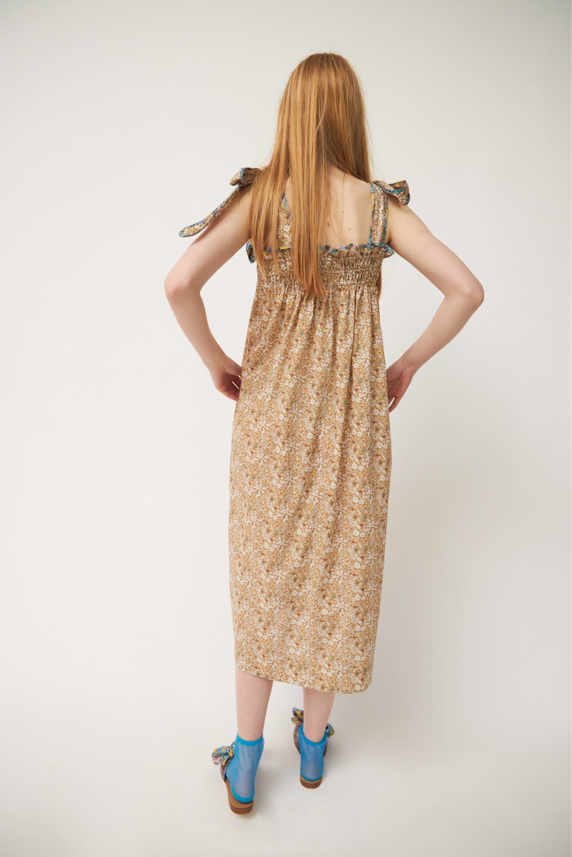 Woman in strap dress with beige and brown flower pattern and shoulder bows, blue socks and sandals with flower patterned bows