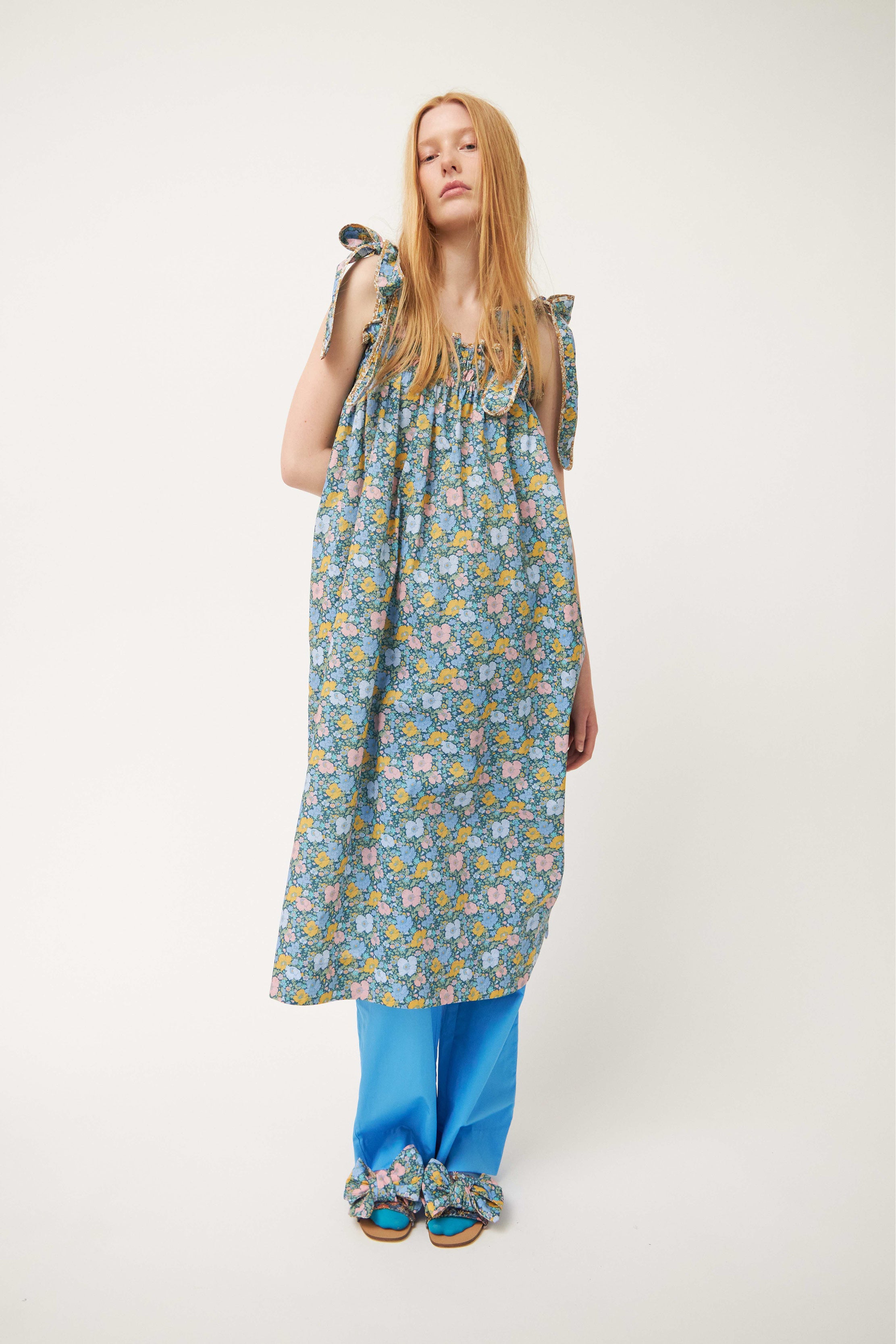 Woman in strap dress with green and multi colored flower pattern and shoulder bows, blue trousers and sandals with bows