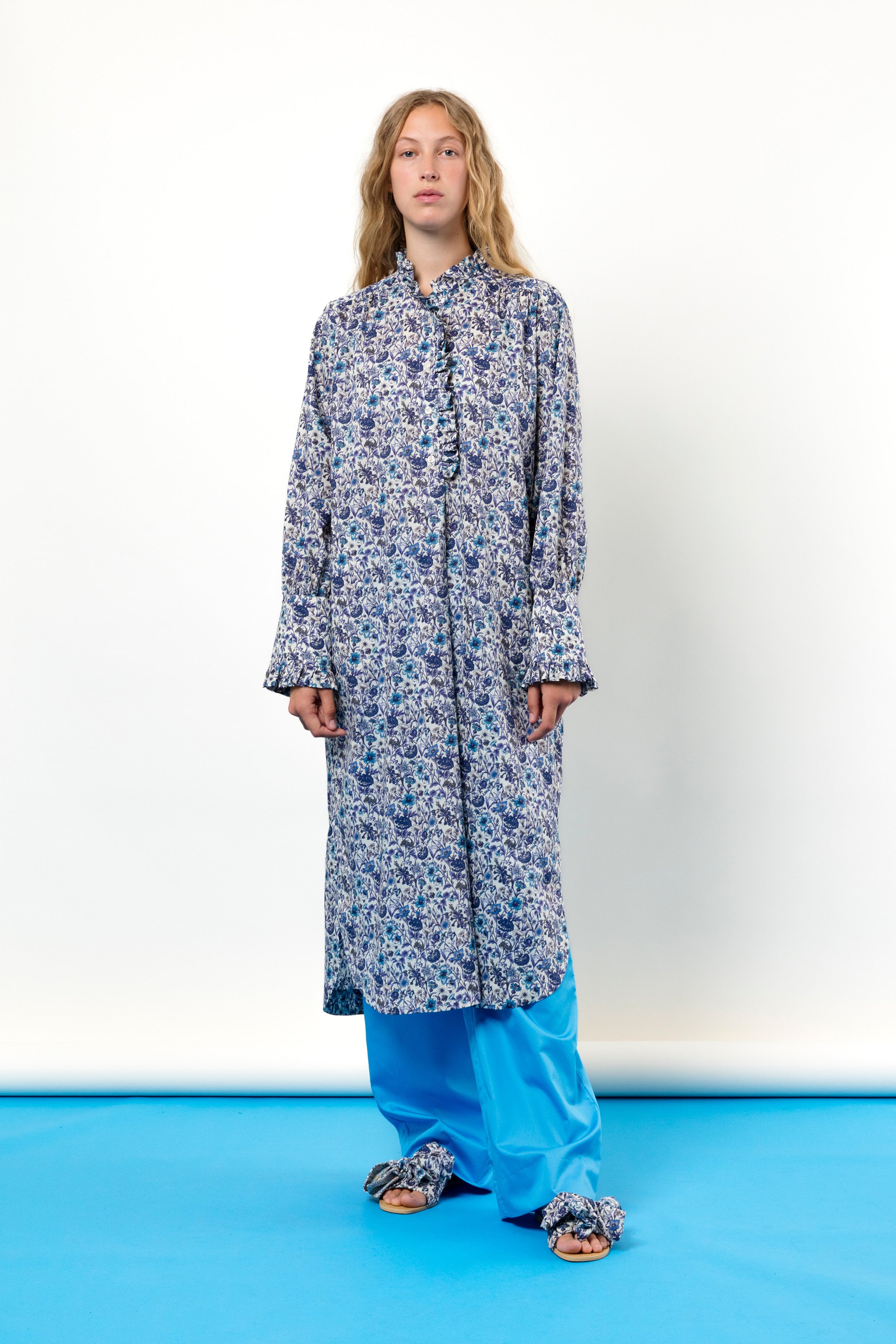 Woman in blue tunic dress with flower pattern, buttons and frills, blue pants and sandals with bows
