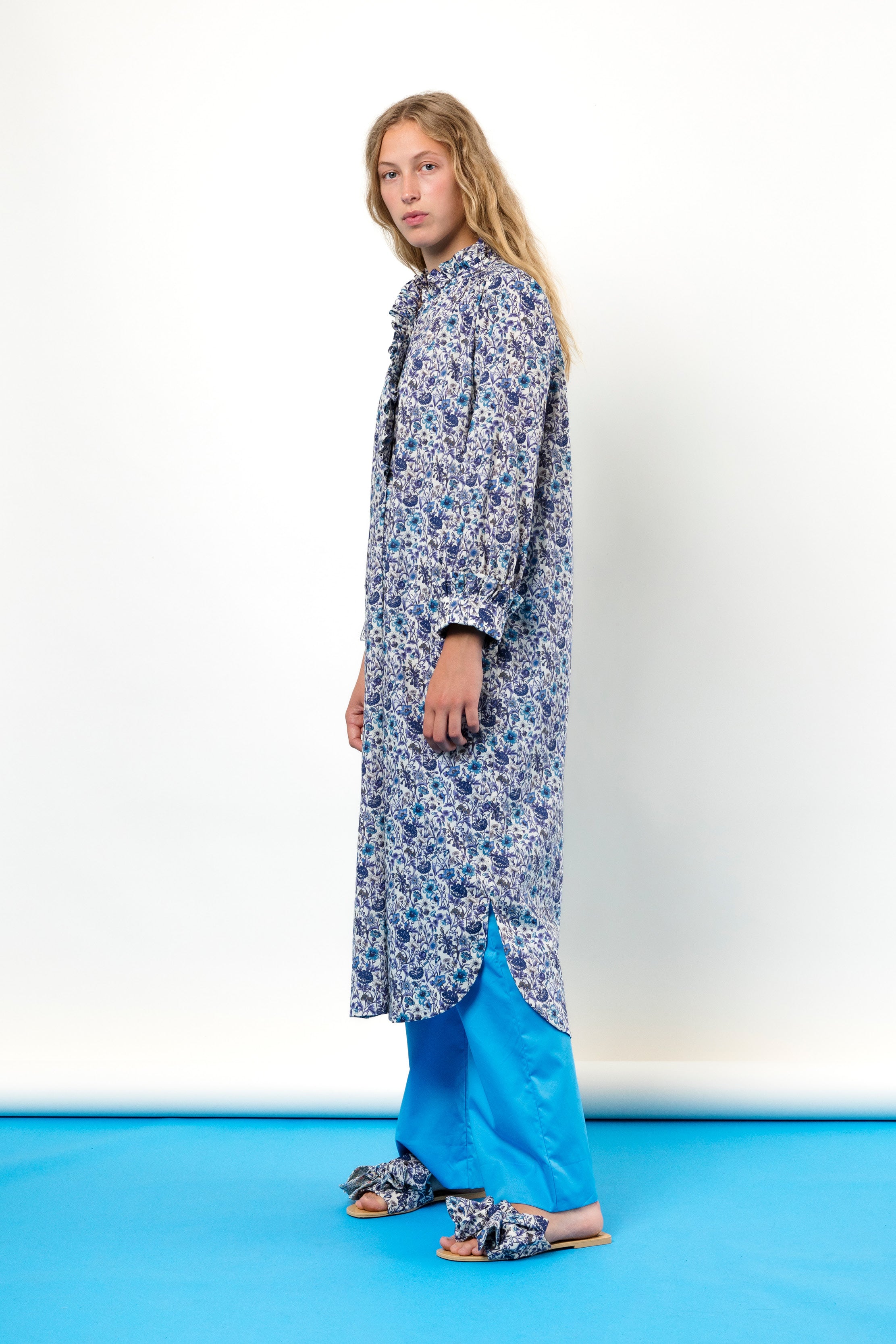 Woman in blue tunic dress with flower pattern, buttons and frills, blue pants and sandals with bows
