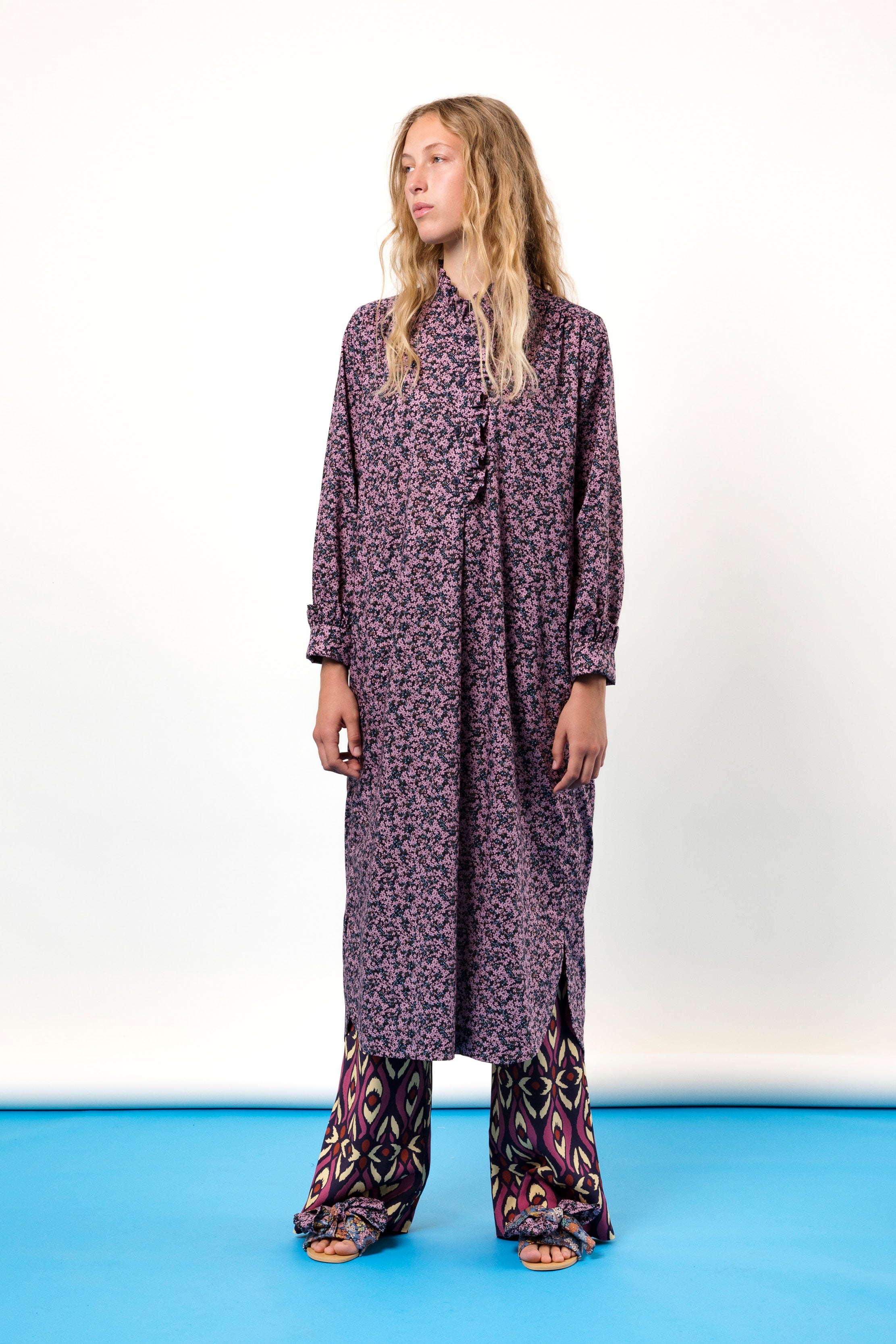 Woman in purple tunic dress with flower pattern, buttons and frills, pants and sandals with bows