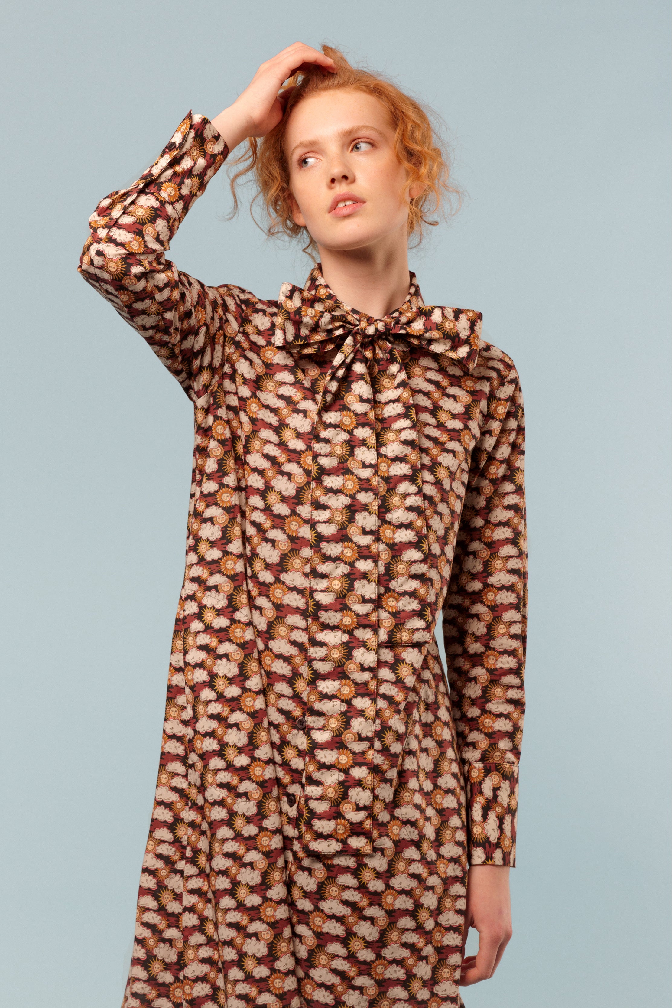 Woman wearing cotton shirt dress with sun and clouds pattern, long sleeves, bow around neck