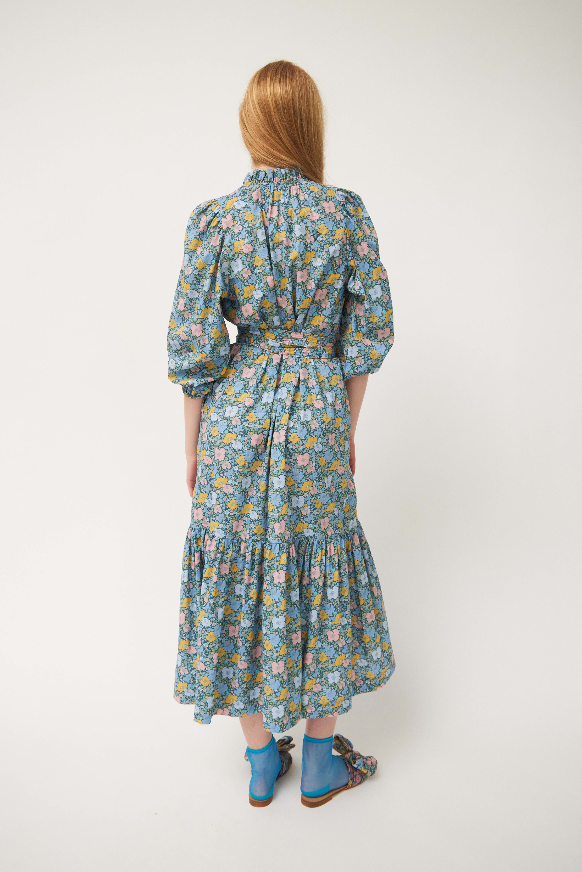 Woman in long dress with multi-colored flower pattern, puffed long sleeves and belt, blue socks and sandals with flower patterned sandals