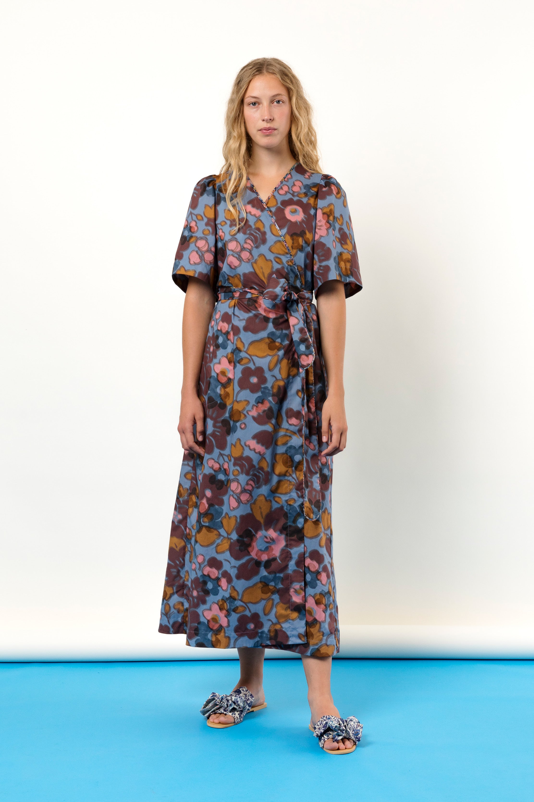 Woman in blue pink and burgundy flower patterned wrap dress in Liberty cotton with short sleeves and sandals with bows