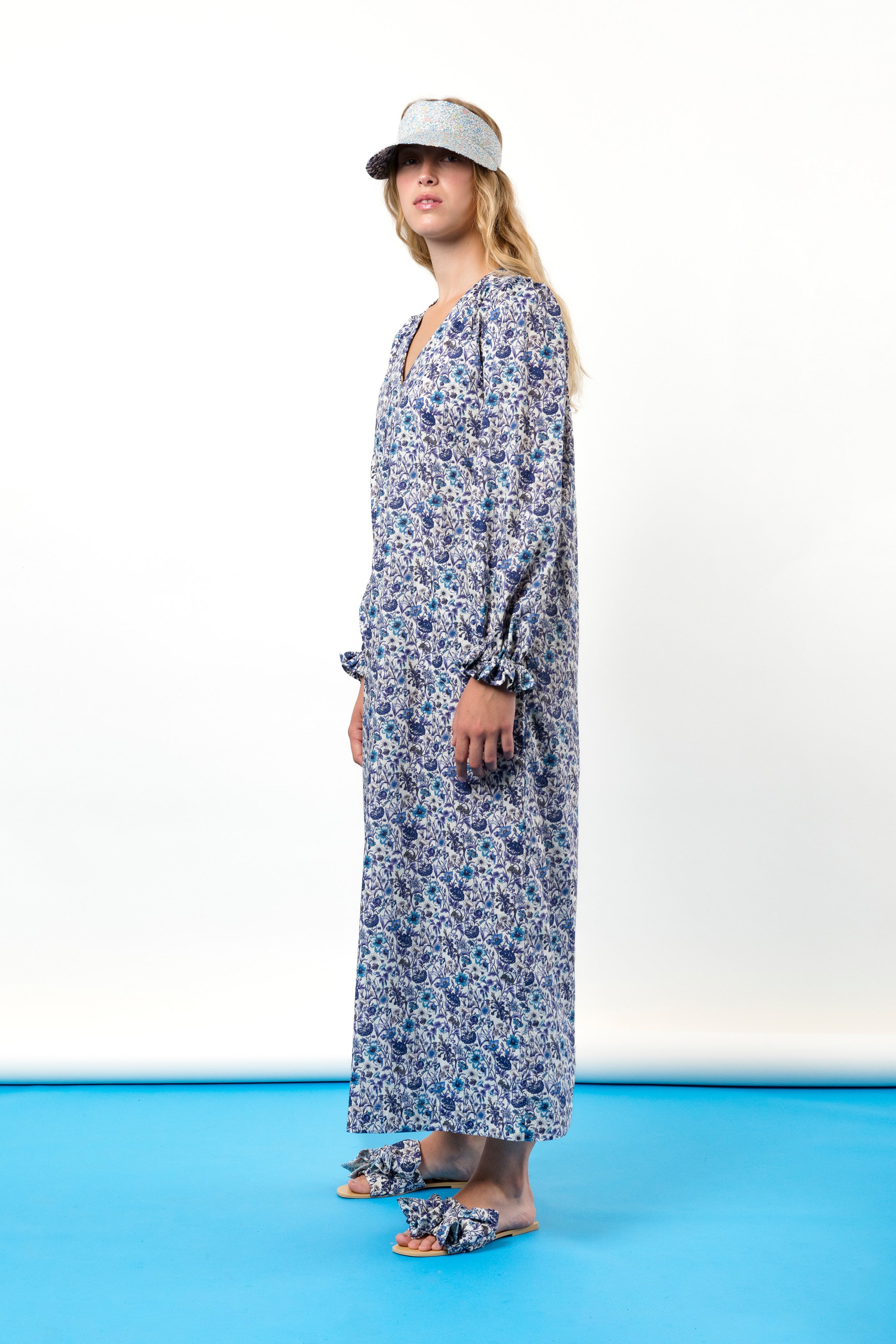 Woman in midi cotton dress with blue flower print, v-neck and ruffled cuffs, sun shade cap and sandals with bows