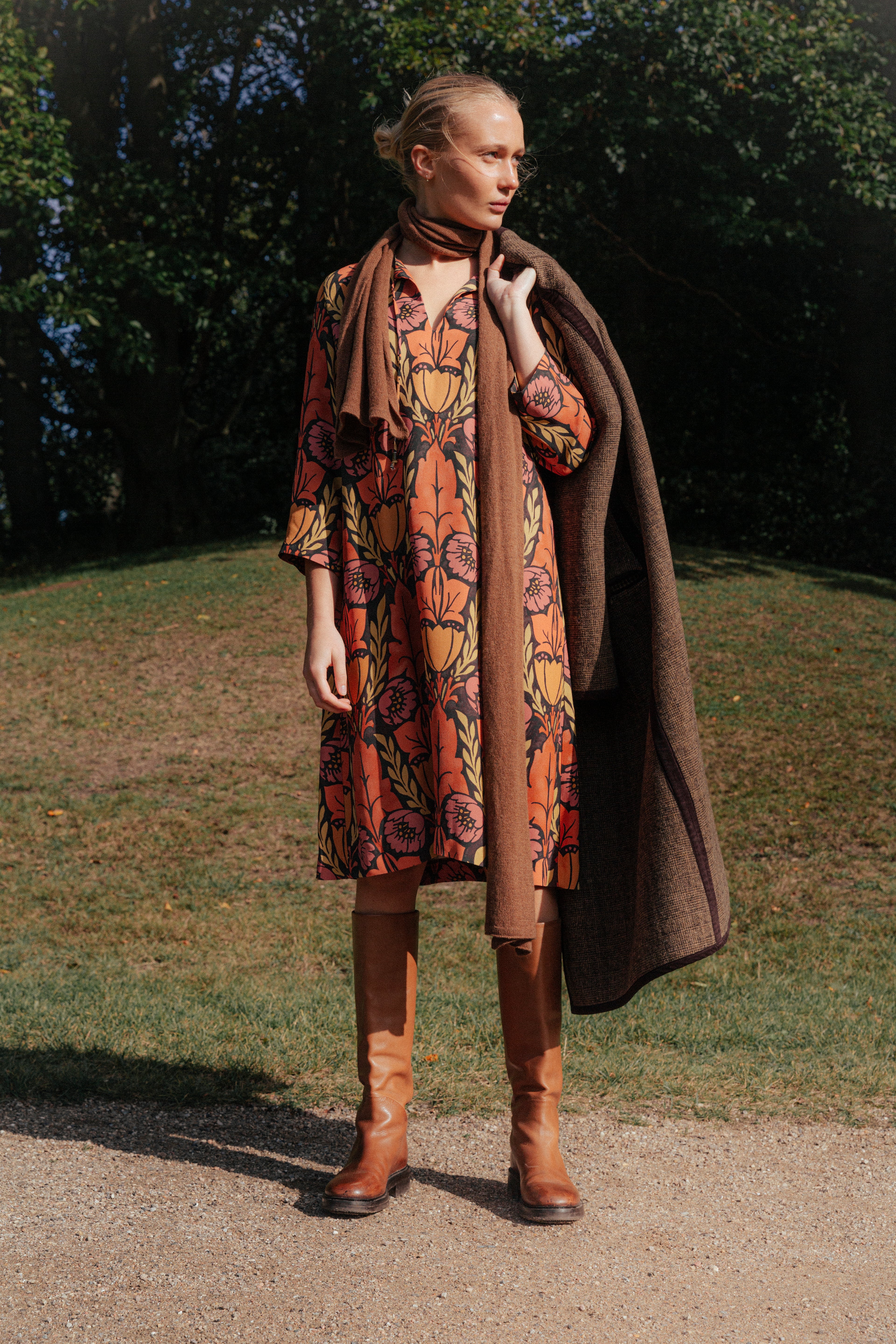 Woman in midi long sleeved viscose dress with large flower print in pink, orange and yellow, brown coat, scarf and boots