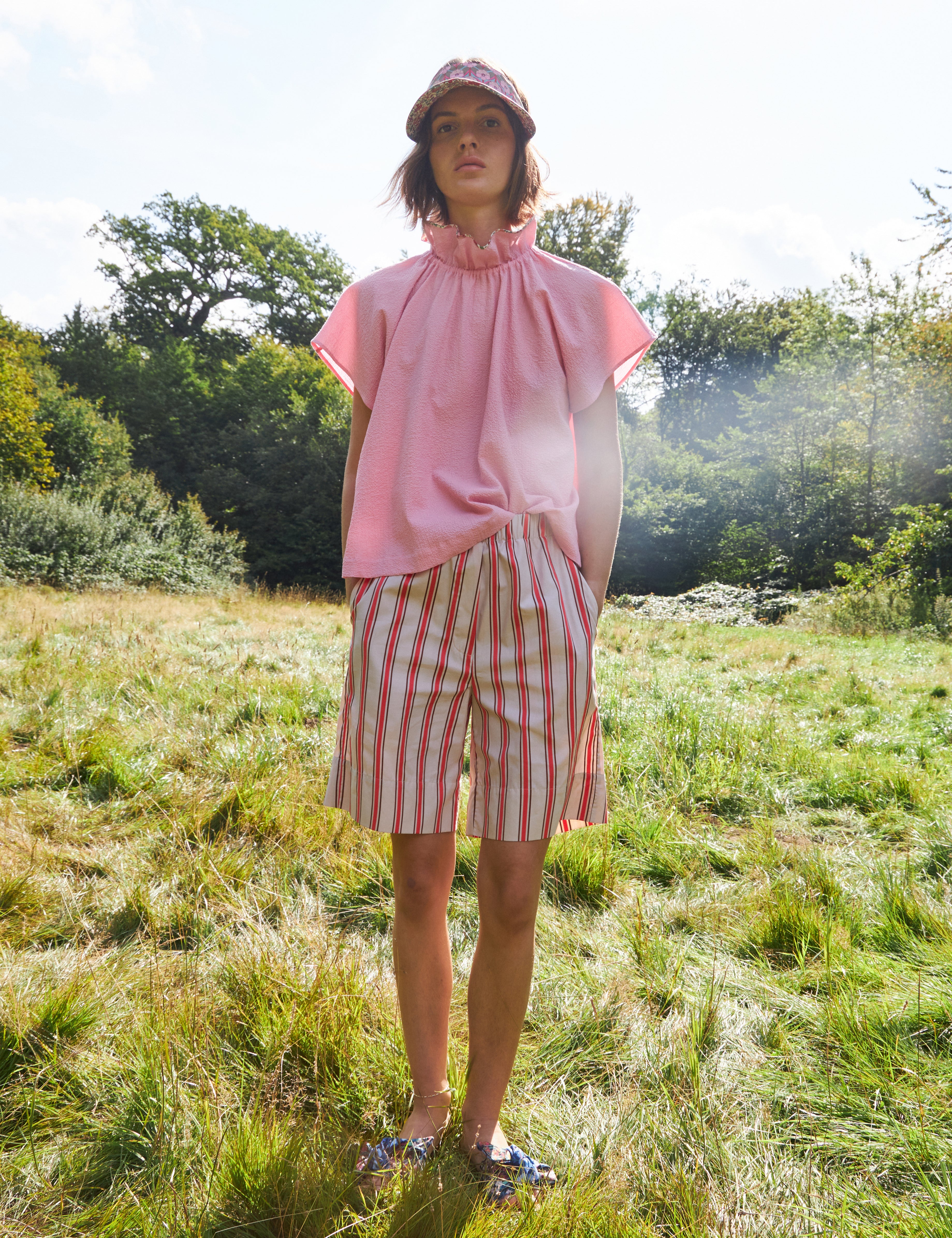 Woman in pink sleeveless top with stand collar and red and white striped shorts and sun cap
