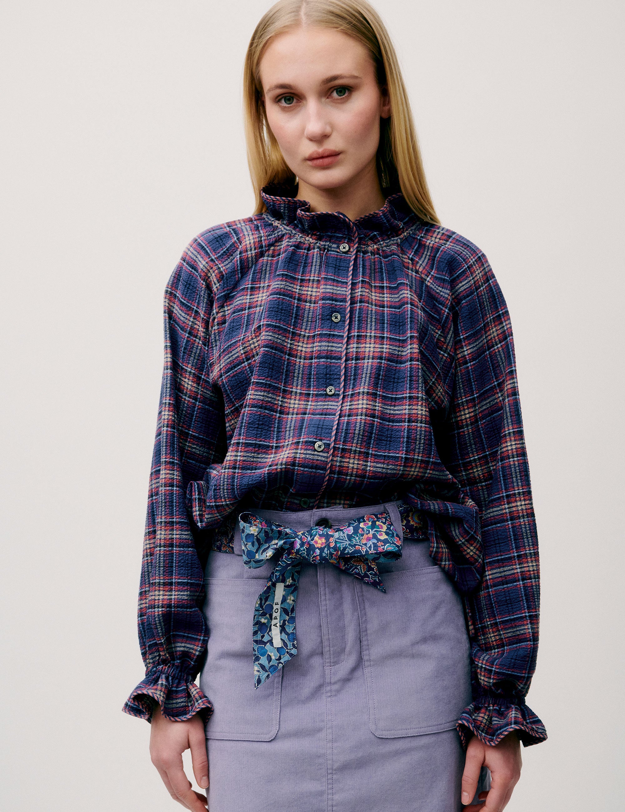 Woman facing forward wearing a dark blue and red checked blouse with a lilac skirt, styled with a floral scarf belt.