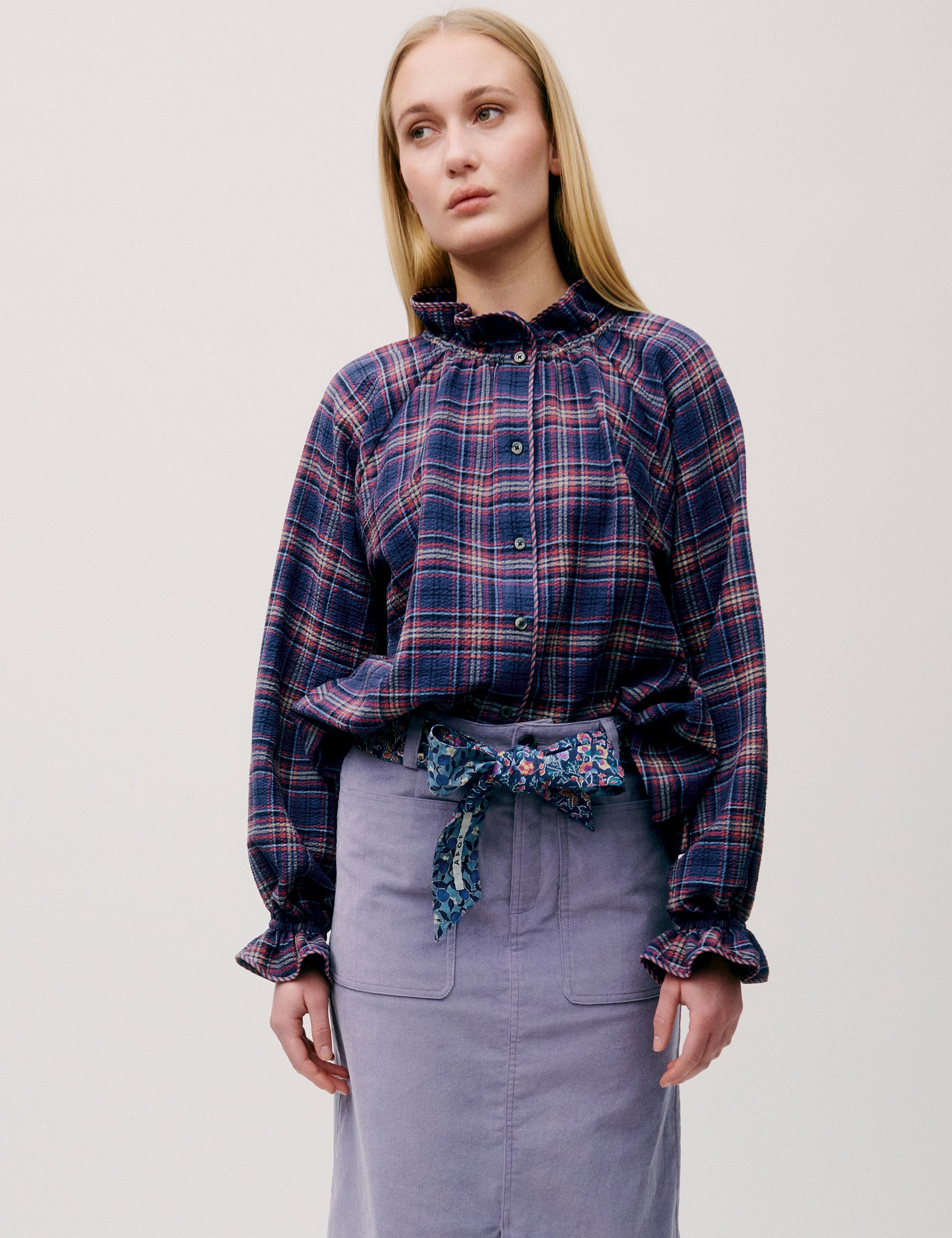 Woman facing forward wearing a dark blue and red checked blouse with a lilac skirt, styled with a floral scarf belt.