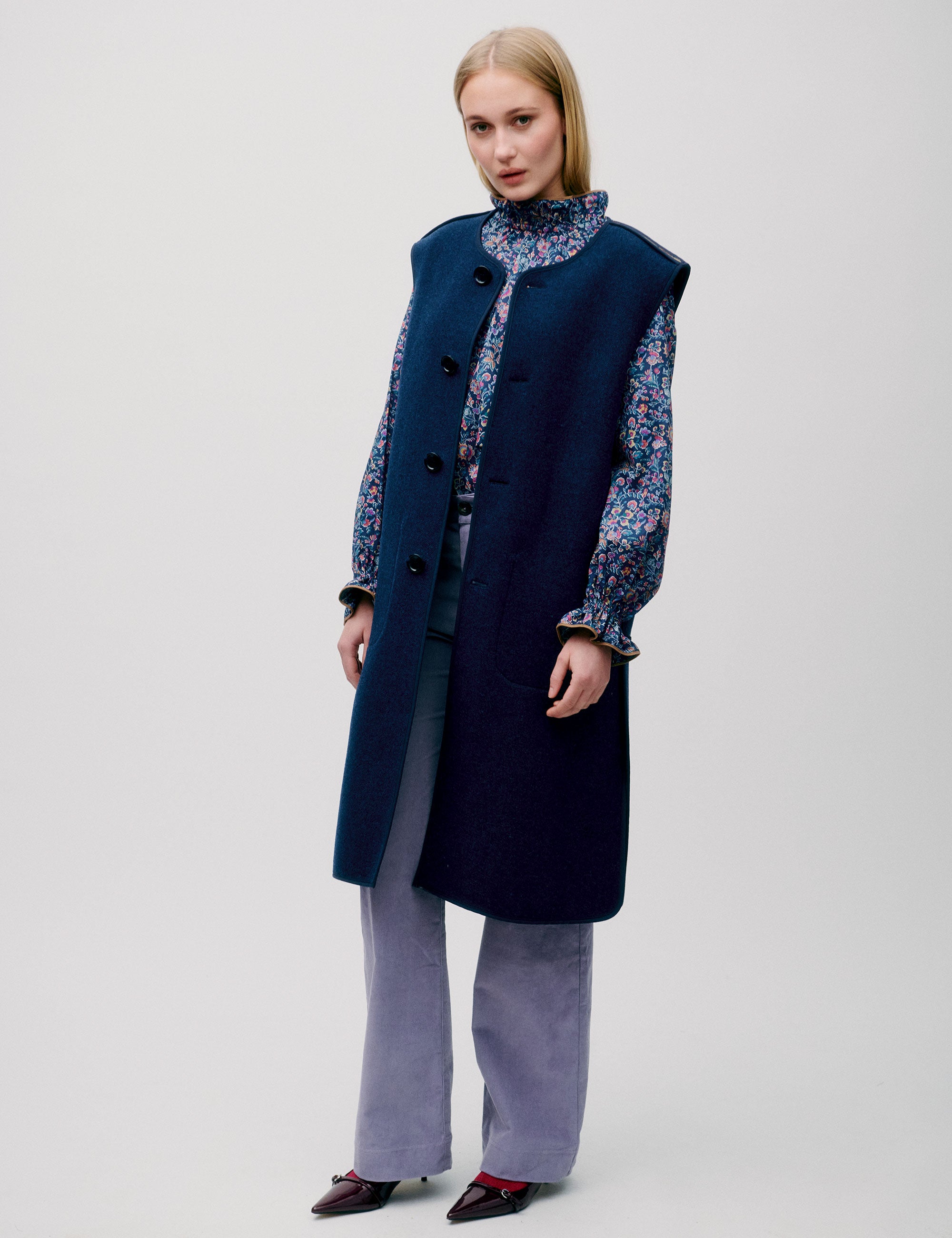 Woman in lavender colored pants, blue flower patterned blouse with frills and long, dark blue sleeveless wool vest