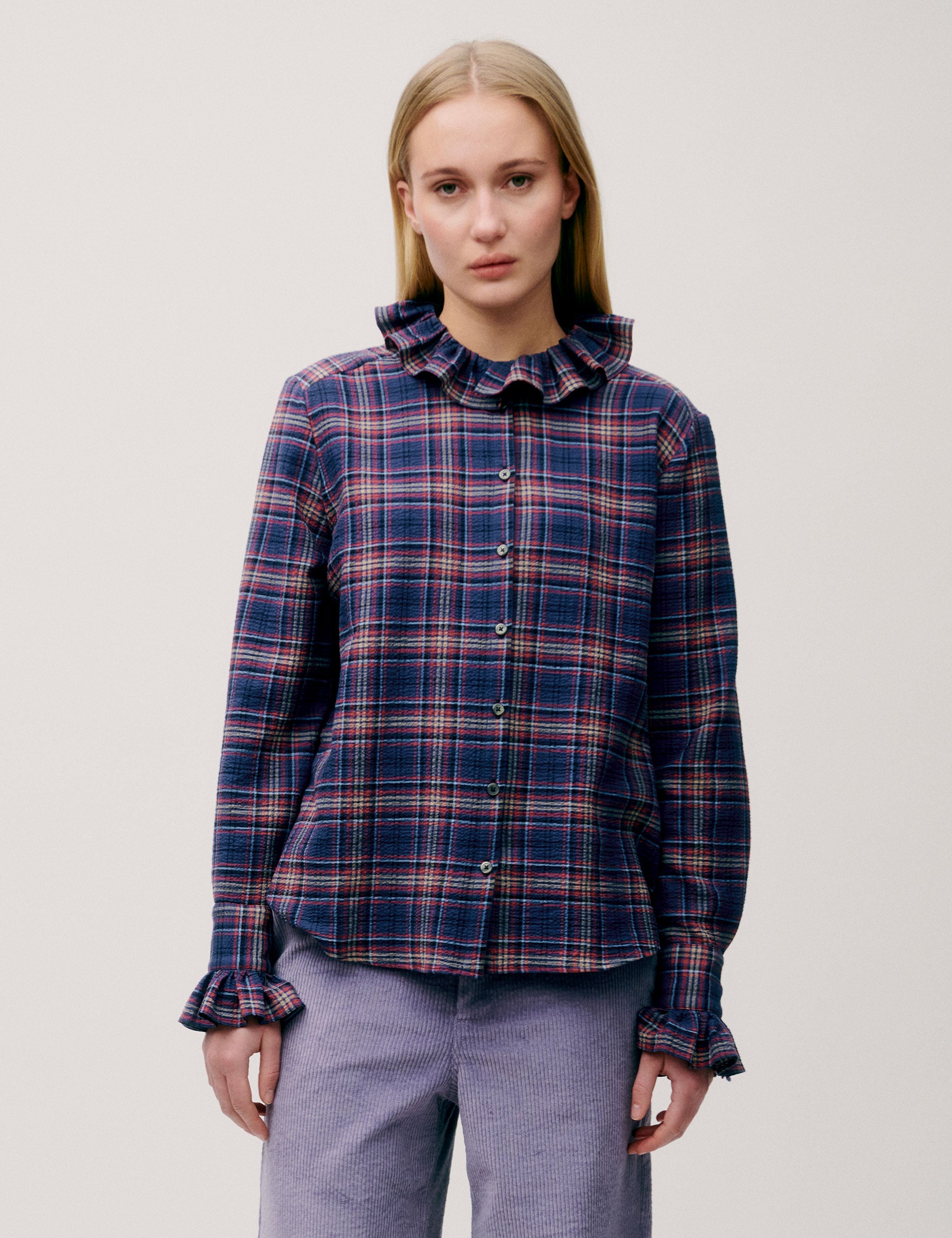 Woman wearing a dark blue and red checked blouse with ruffle collar and cuffs, paired with lilac corduroy trousers