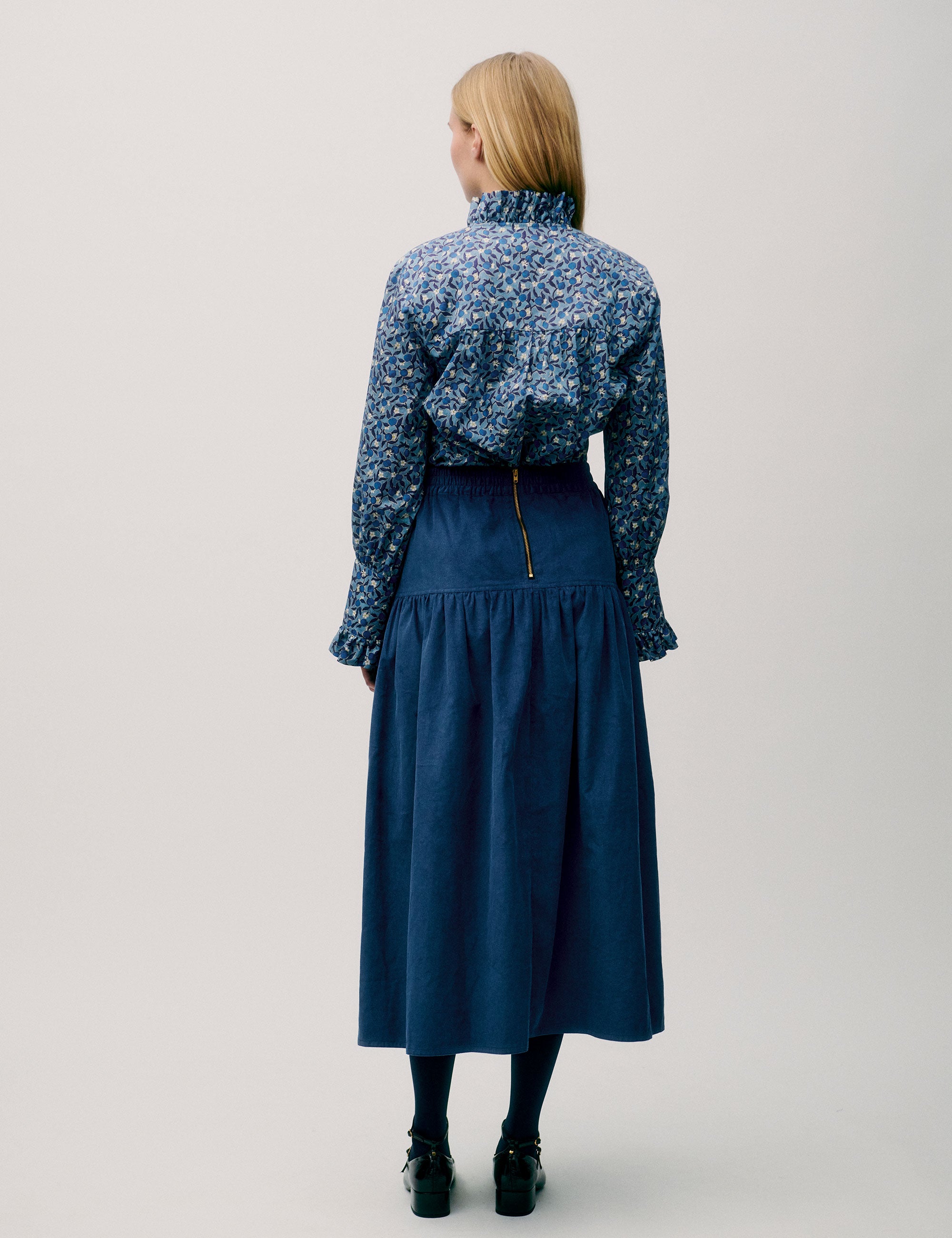 Woman in long blue corduroy skirt and blue floral long sleeved blouse with ruffled neck and cuffs