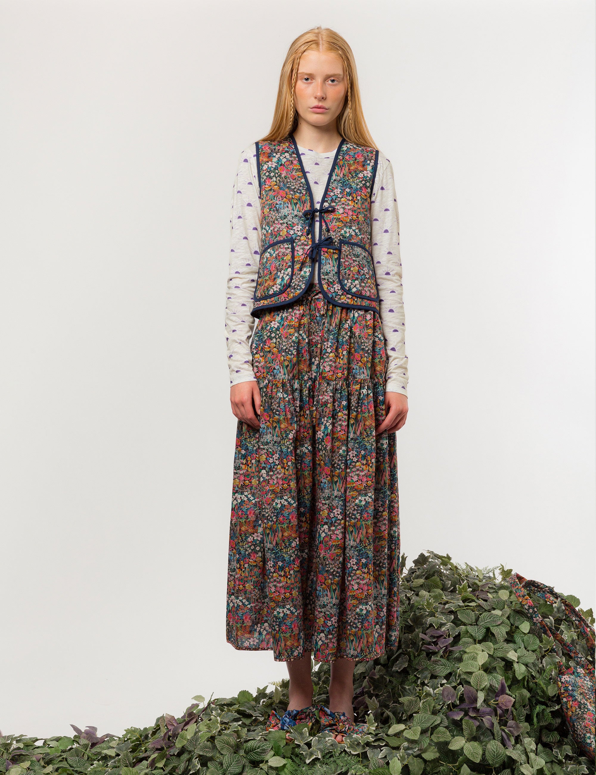 Woman in reversible liberty cotton multi-colored floral print vest with buttons, pockets and blue pipings, long skirt with floral print and long sleeved white t-shirt with sunset print