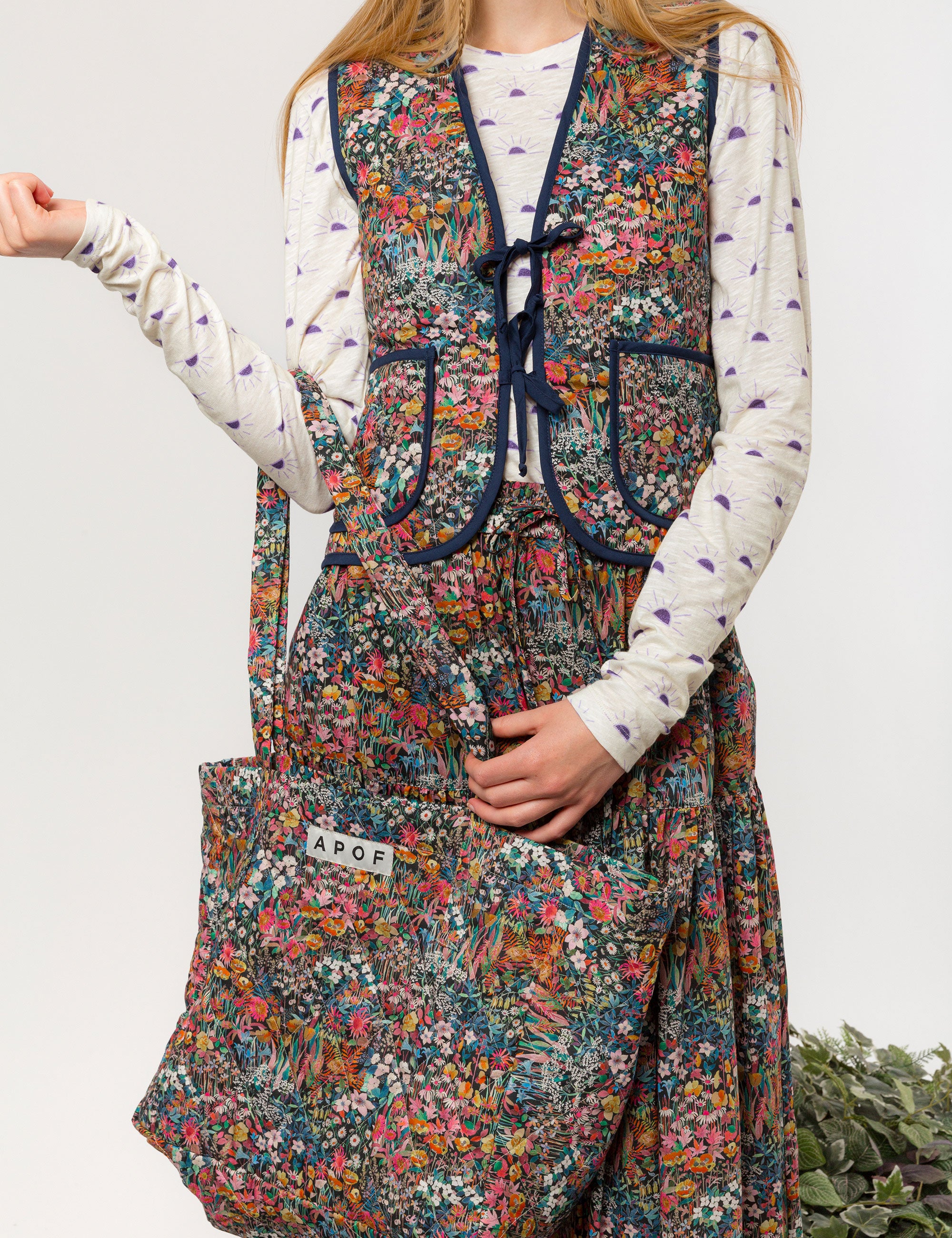 Woman in reversible liberty cotton multi-colored floral print vest with buttons, pockets and blue pipings, long skirt with floral print and long sleeved white t-shirt with sunset print