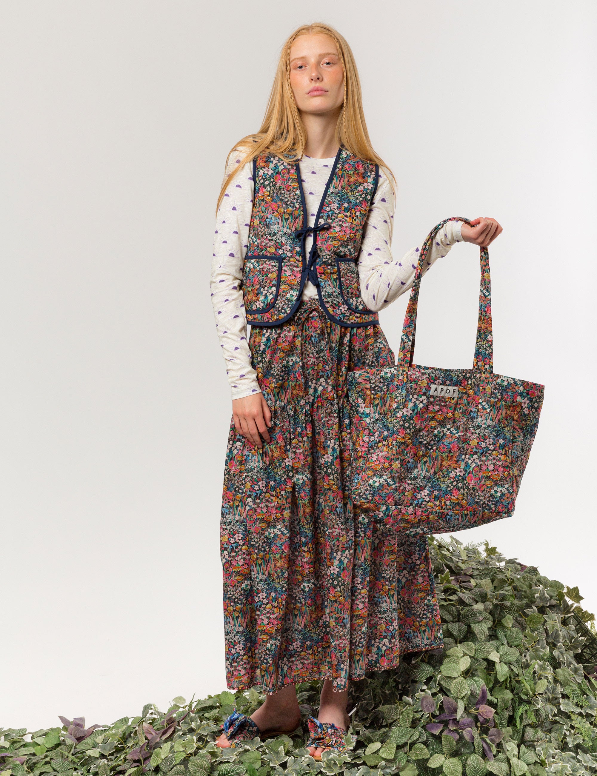 Woman in reversible liberty cotton multi-colored floral print vest with buttons, pockets and blue pipings, long skirt with floral print and long sleeved white t-shirt with sunset print, carrying an APOF bag
