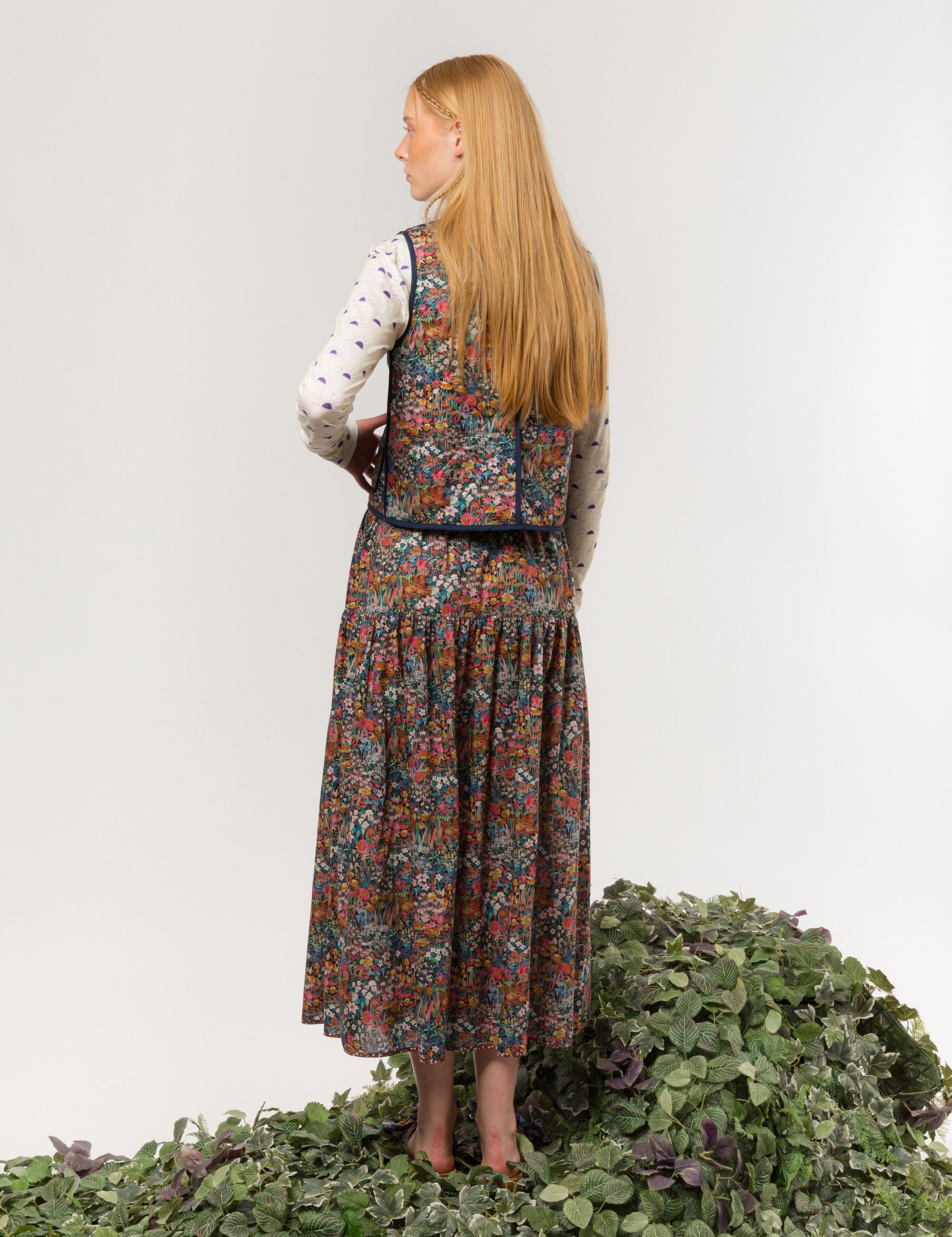 Woman in reversible liberty cotton multi-colored floral print vest with buttons, pockets and blue pipings, long skirt with floral print and long sleeved white t-shirt with sunset print