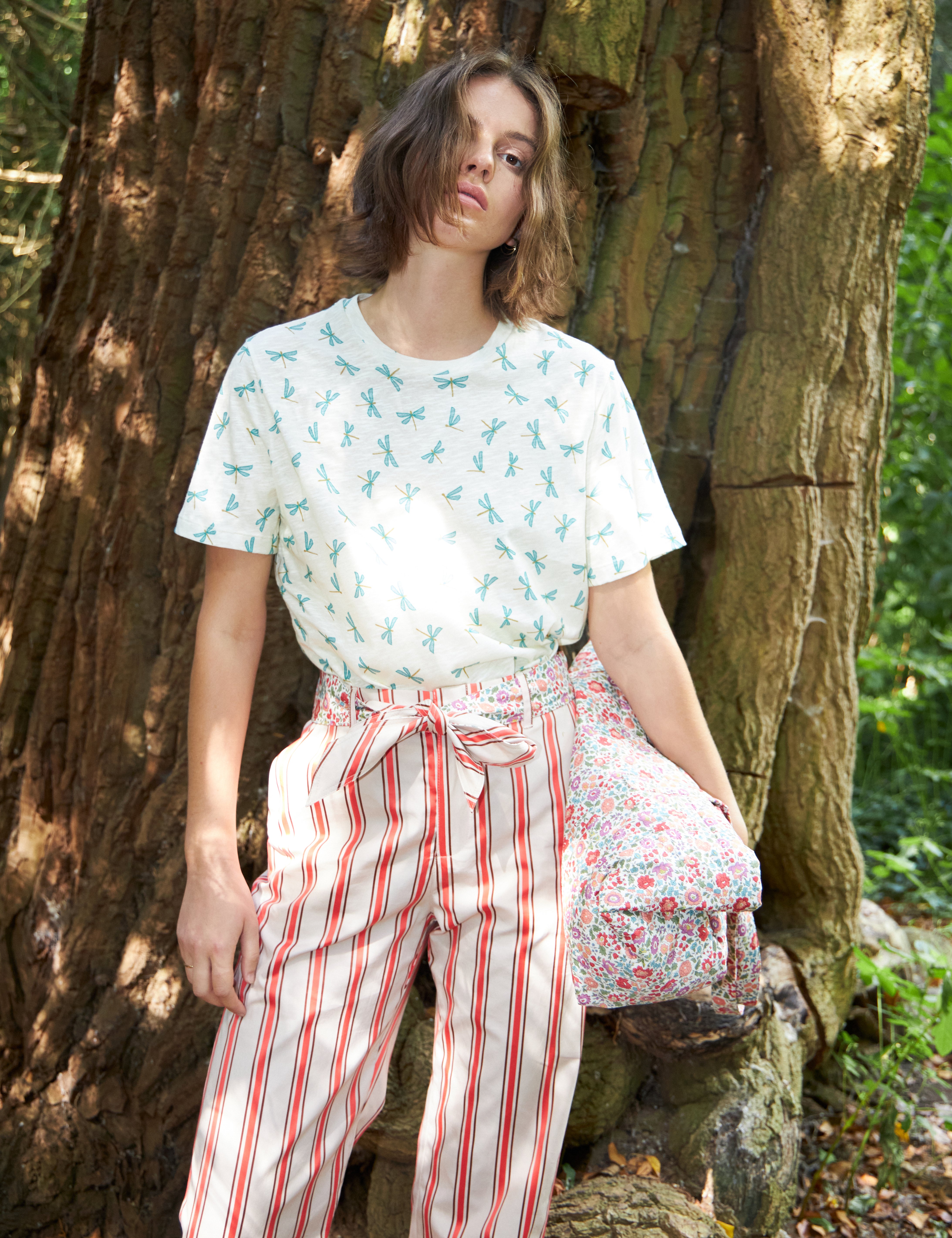 Kvinna i rödvit randiga byxor med bomullsbälte och vit t-shirt med grönt trollsländemönster som bär en flerfärgad blommig bomullsväska