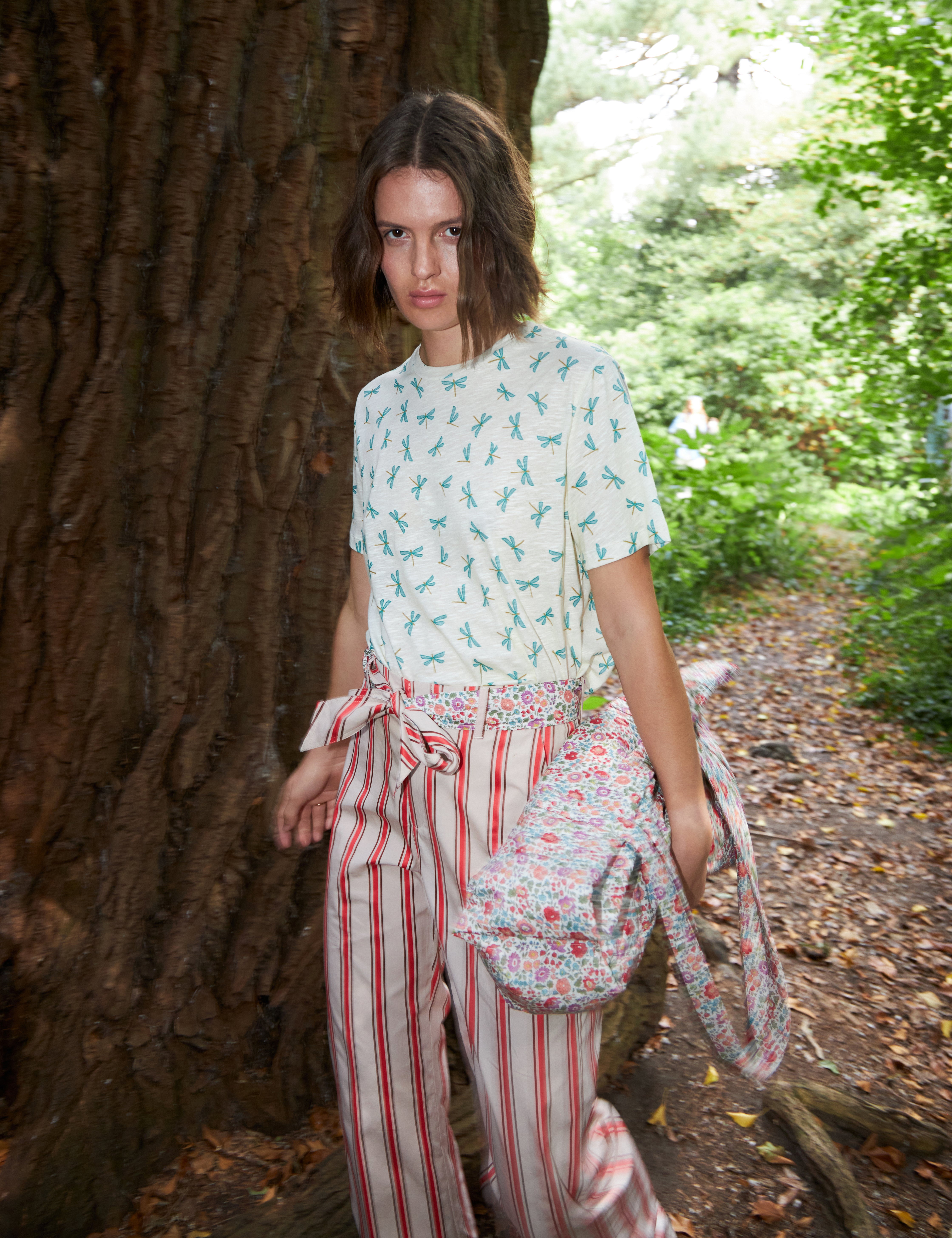 Woman in red and white strped pants with cotton belt and white t-shirt with green dragonfly pattern carrying a multi-colored floral print cotton tote bag