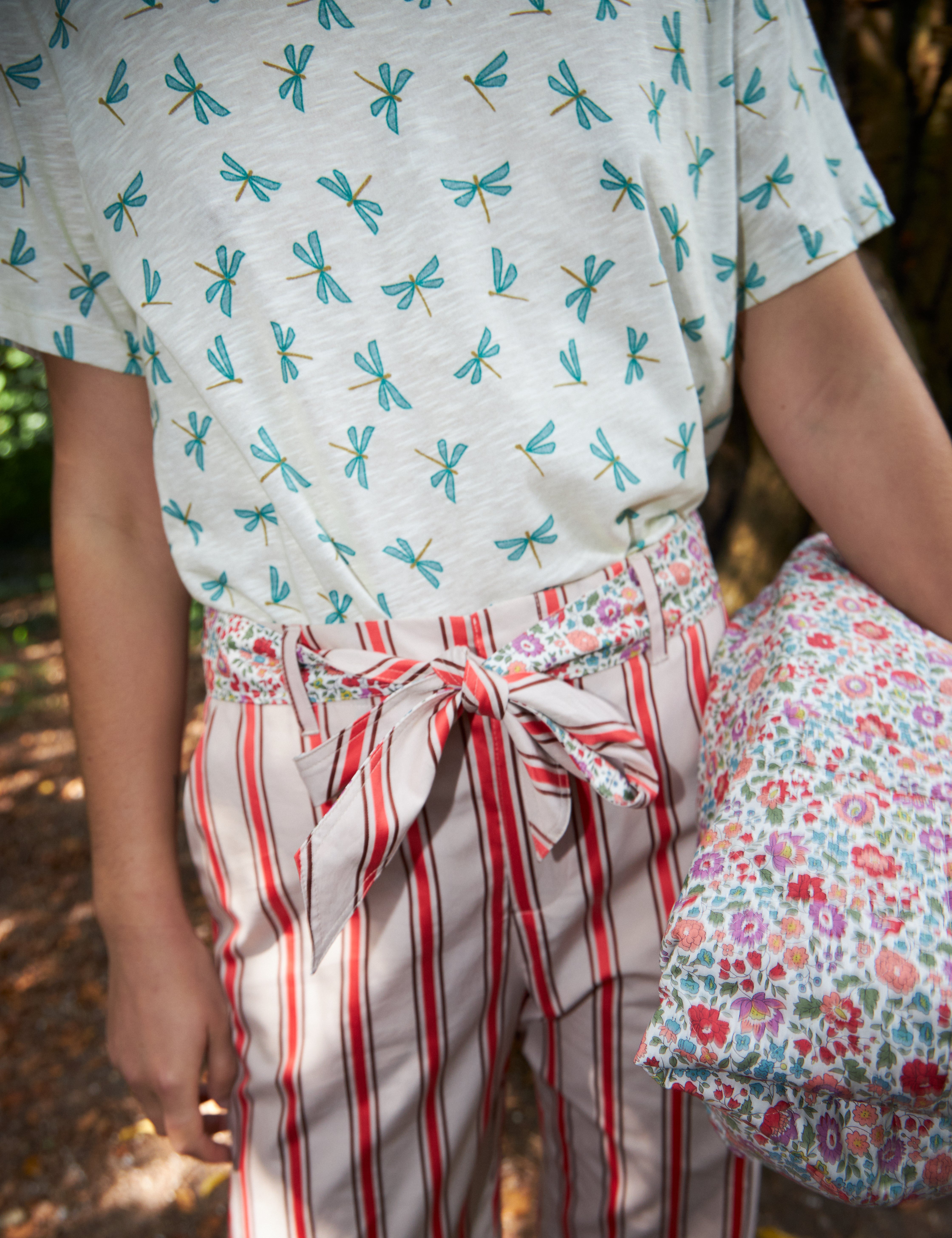 Kvinna i röd- och vitrandiga byxor med bomullsbälte och vit t-shirt med grönt trollsländemönster samt flerfärgad blommig bomullsväska