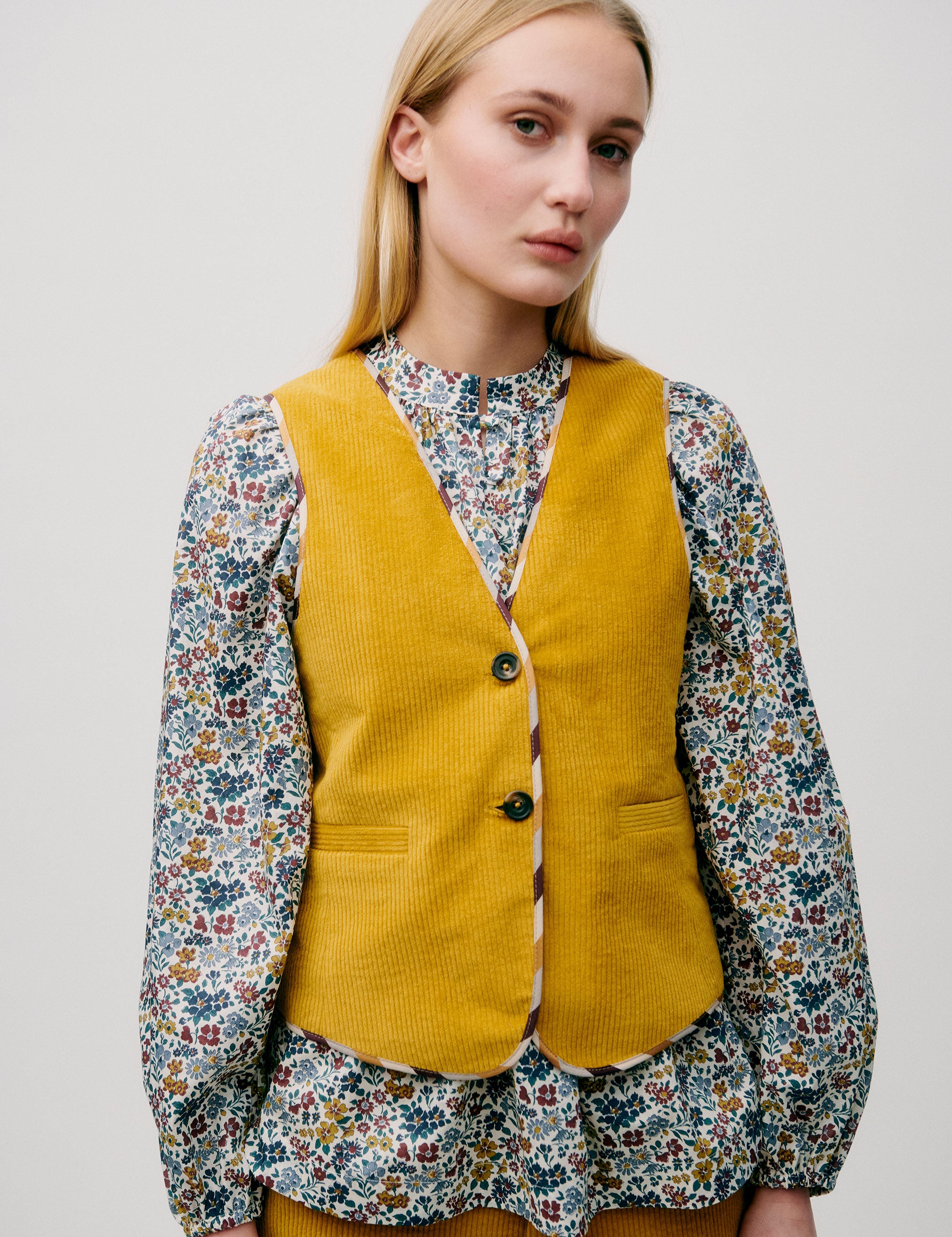 Woman facing forward wearing a yellow corduroy vest layered over a long-sleeved floral blouse and matching trousers.