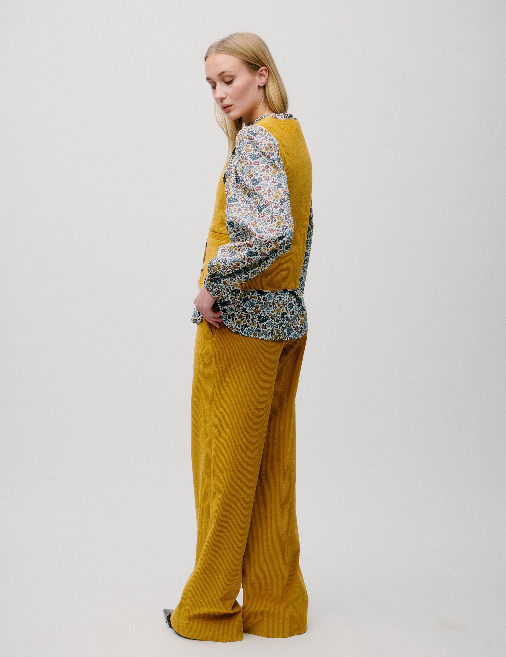 Woman in profile wearing a yellow corduroy vest over a floral blouse with yellow trousers.
