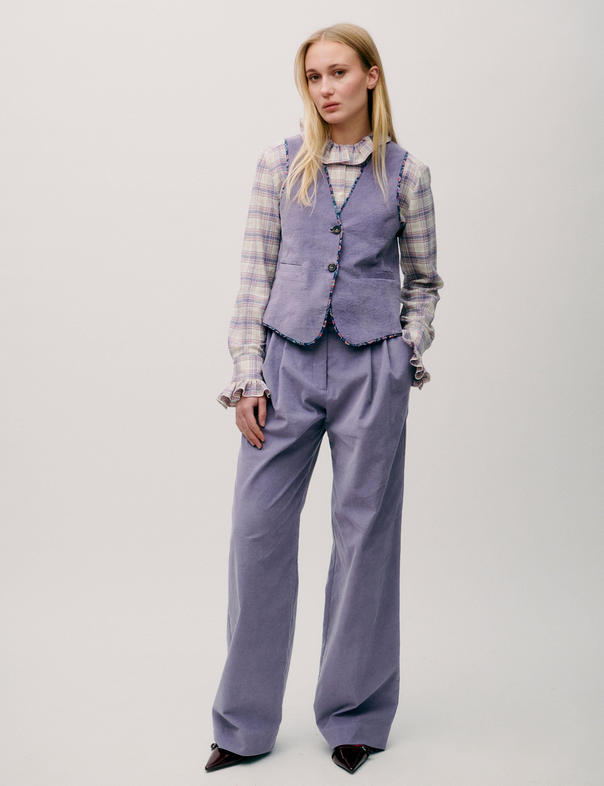 Woman wearing a checked blouse with ruffle collar under a striped lilac vest and high-waisted wide lilac corduroy trousers