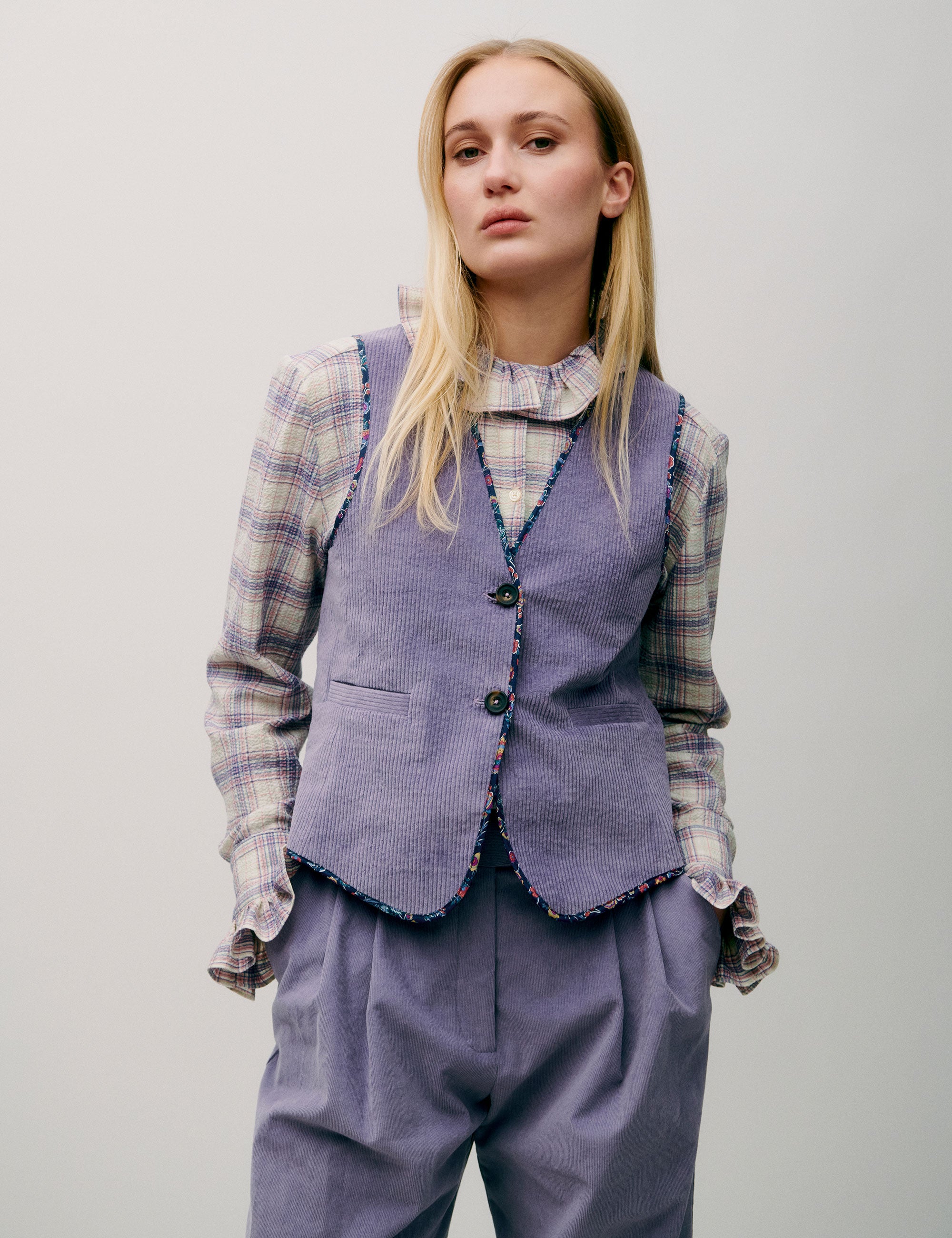 Woman wearing a checked blouse with ruffle collar under a striped lilac vest and high-waisted lilac corduroy trousers
