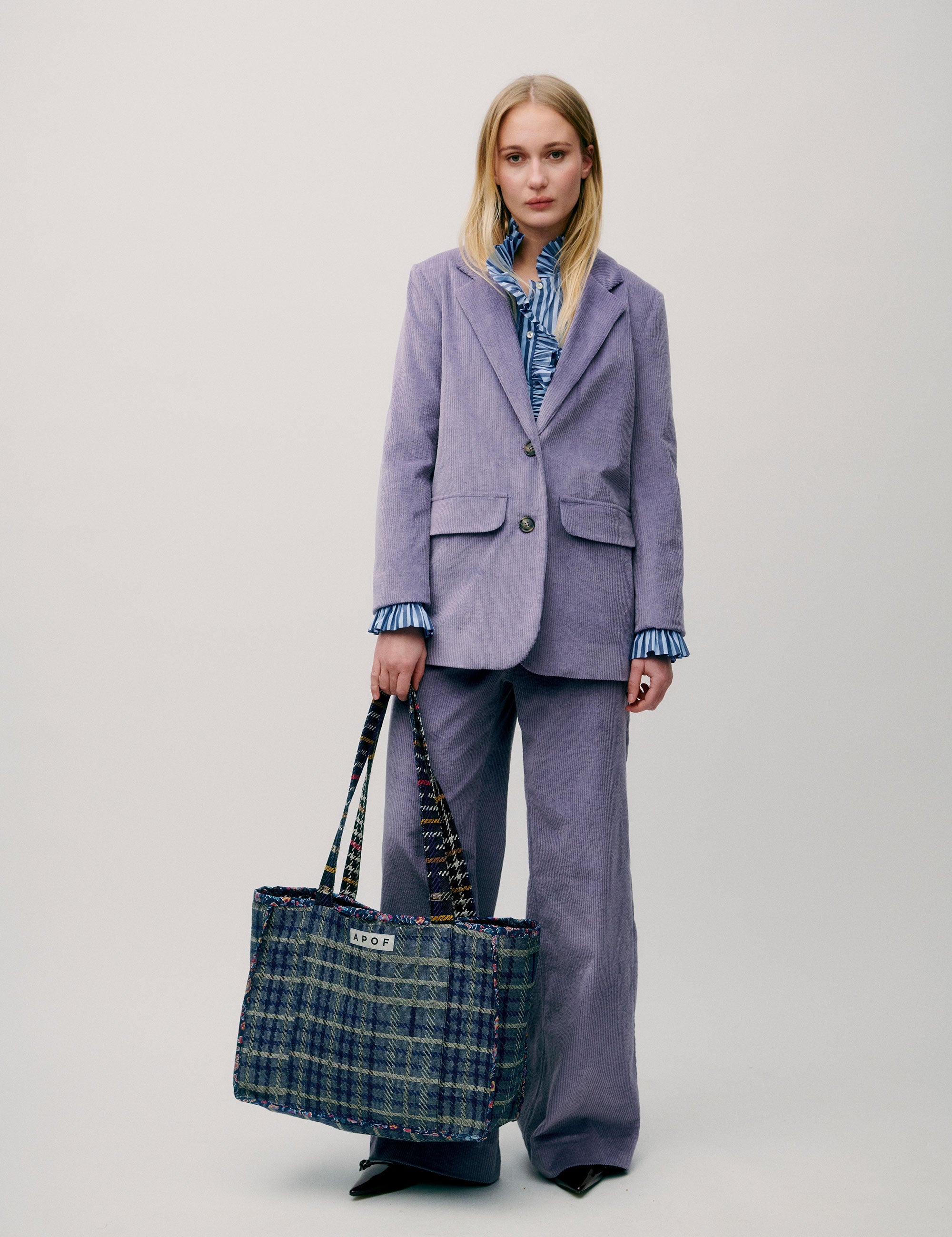 Woman in lavender colored corduroy pants and jacket and blue striped shirt with frills carrying a large check patterned cotton tote bag