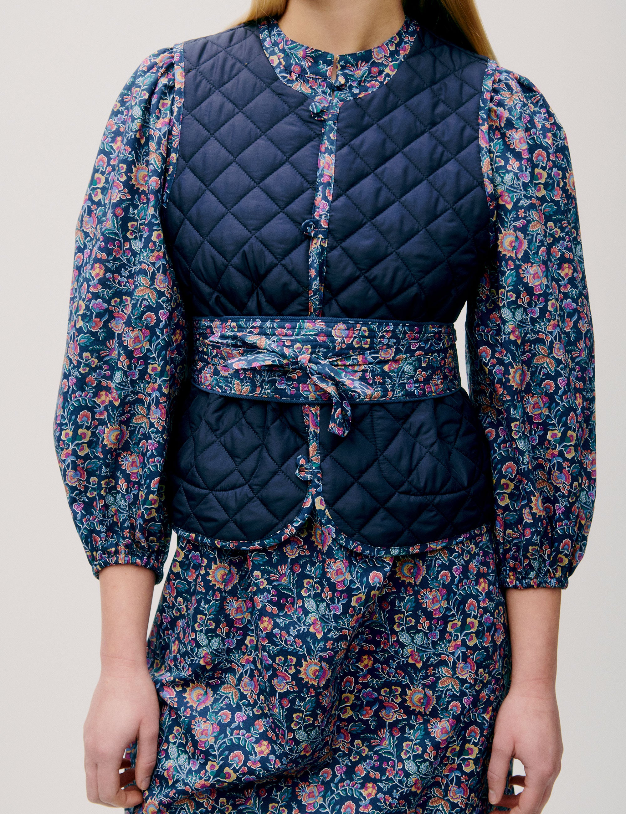 Woman in pink and blue flower patterned short dress with long sleeves, quilted vest and matching cotton belt
