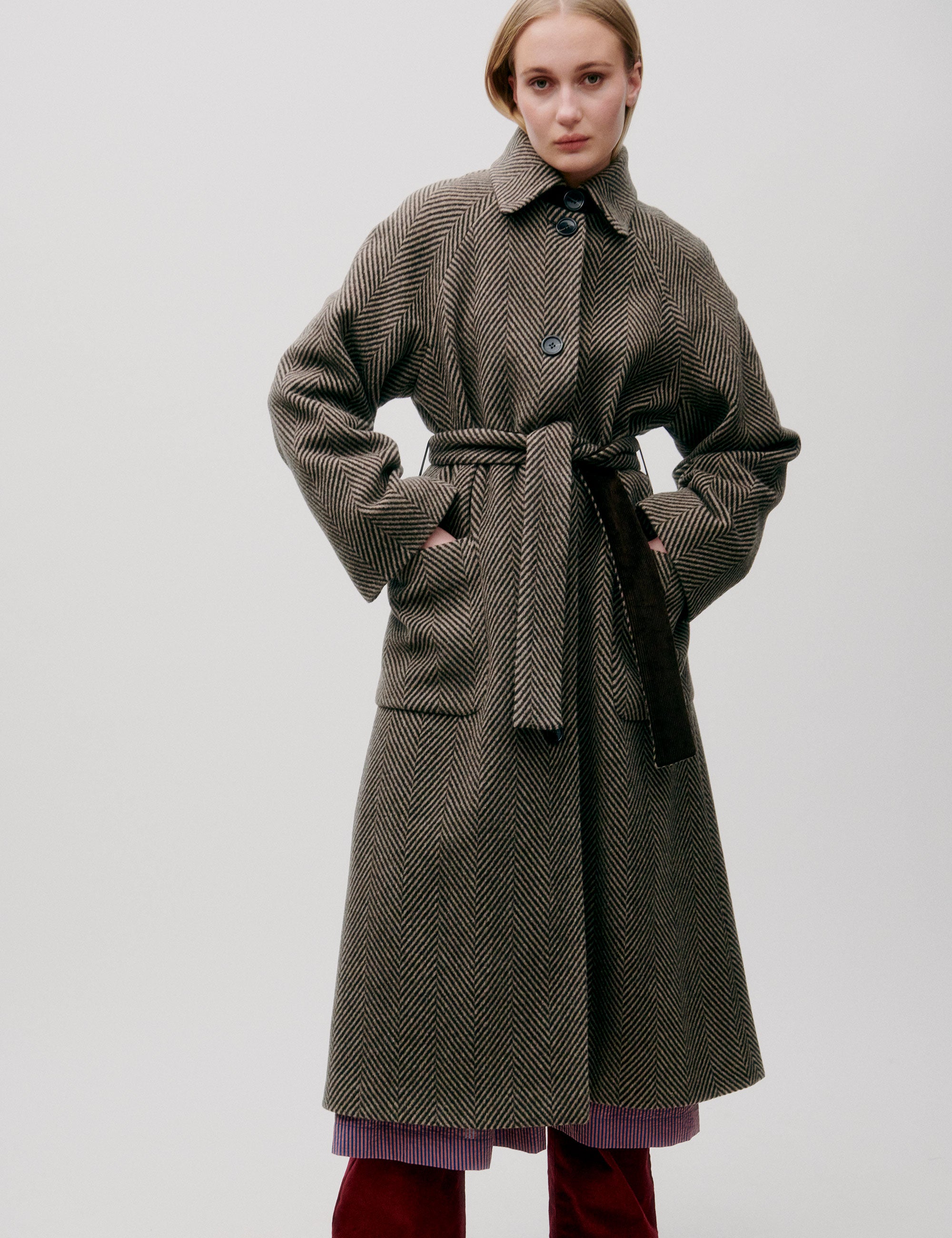 Woman in long herringbone patterned wool coat with collar, pockets, large buttons and matching belt
