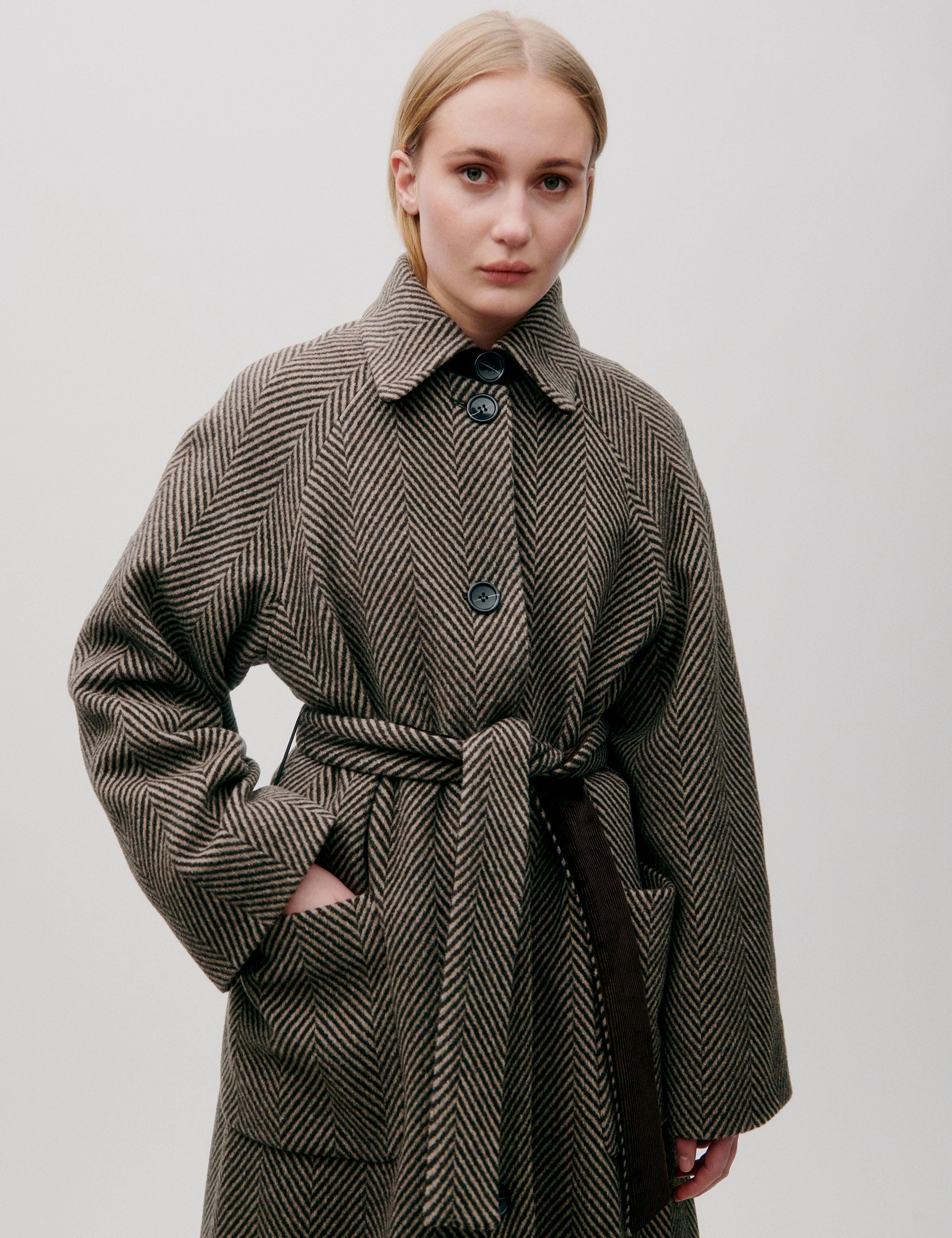 Woman in long herringbone patterned wool coat with collar, pockets, large buttons and matching belt