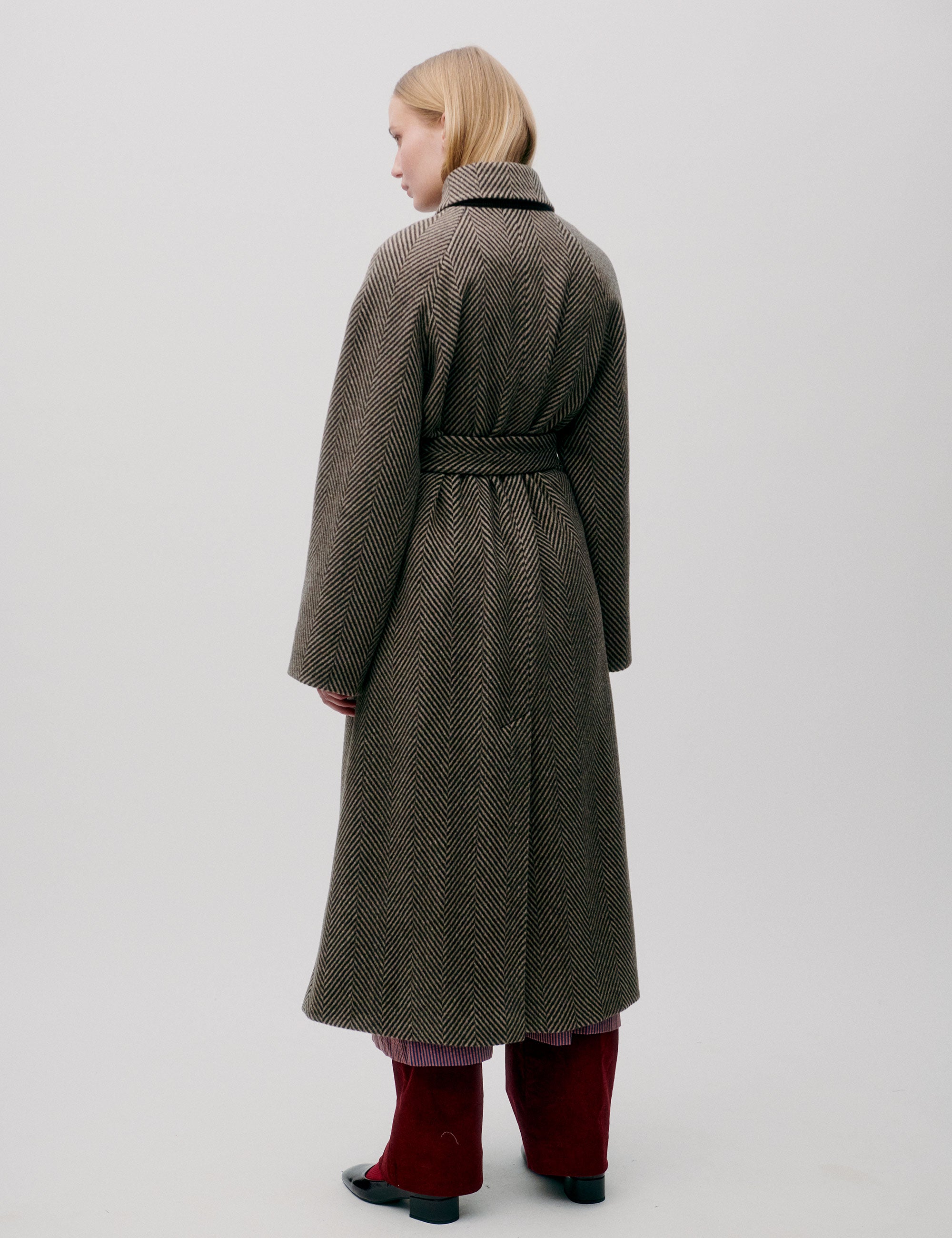 Woman in long herringbone patterned wool coat with collar, pockets, large buttons and matching belt