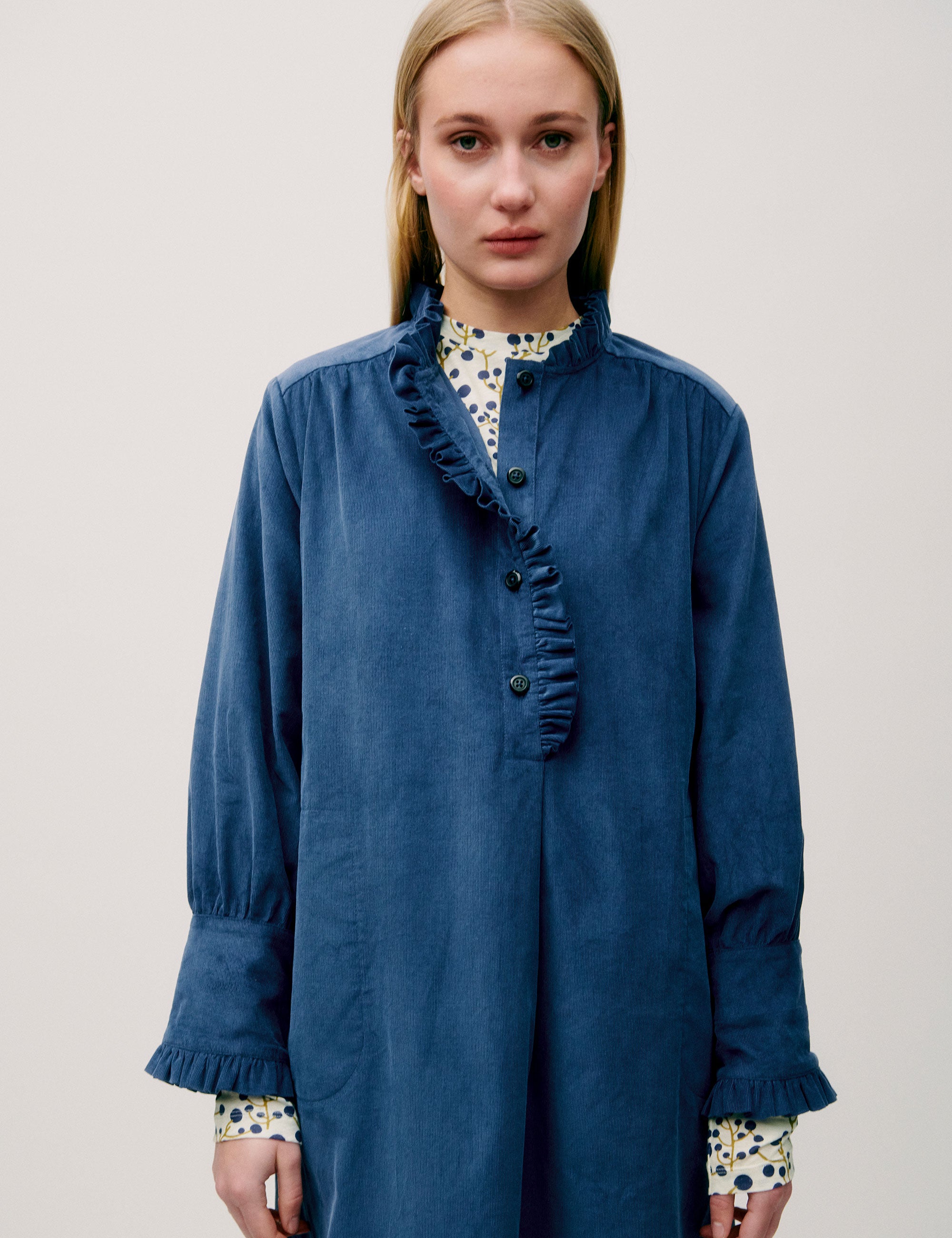 Woman wearing a loose-fitting blue corduroy shirt dress with ruffle detail and floral inner cuffs