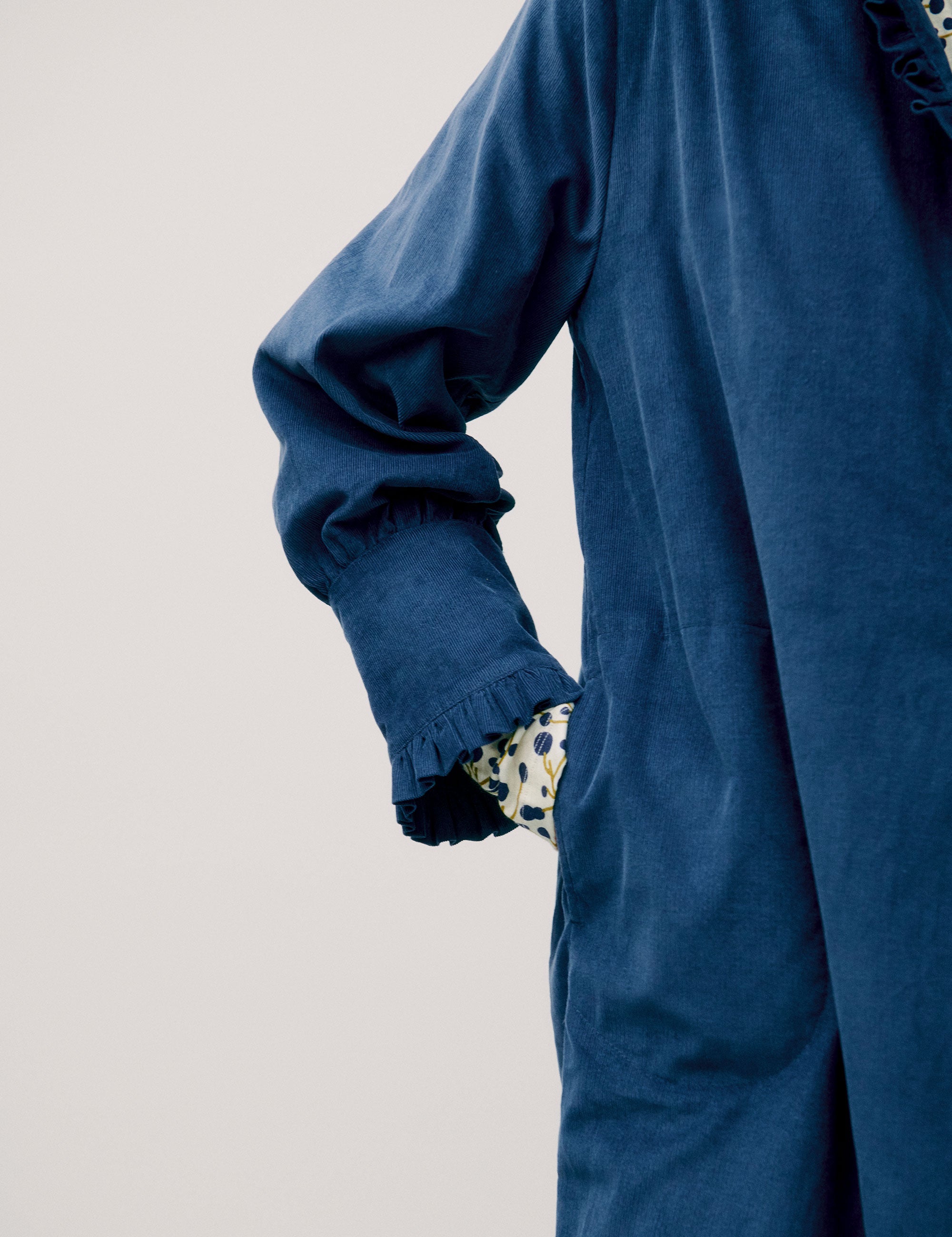 Woman wearing a loose-fitting blue corduroy shirt dress with ruffle detail and floral inner cuffs