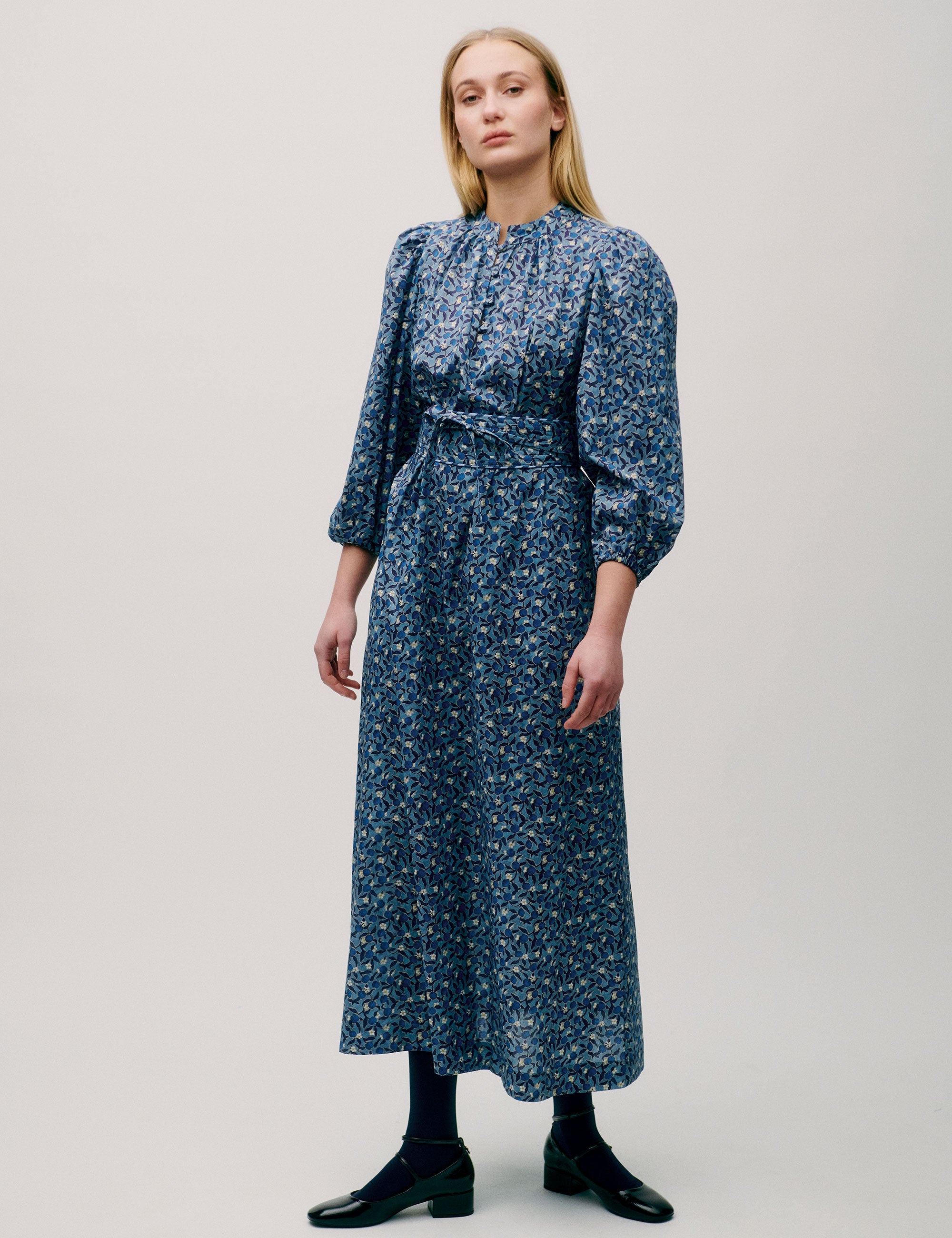 Woman wearing a long blue floral dress with puff sleeves, pleated neckline, and tie waist
