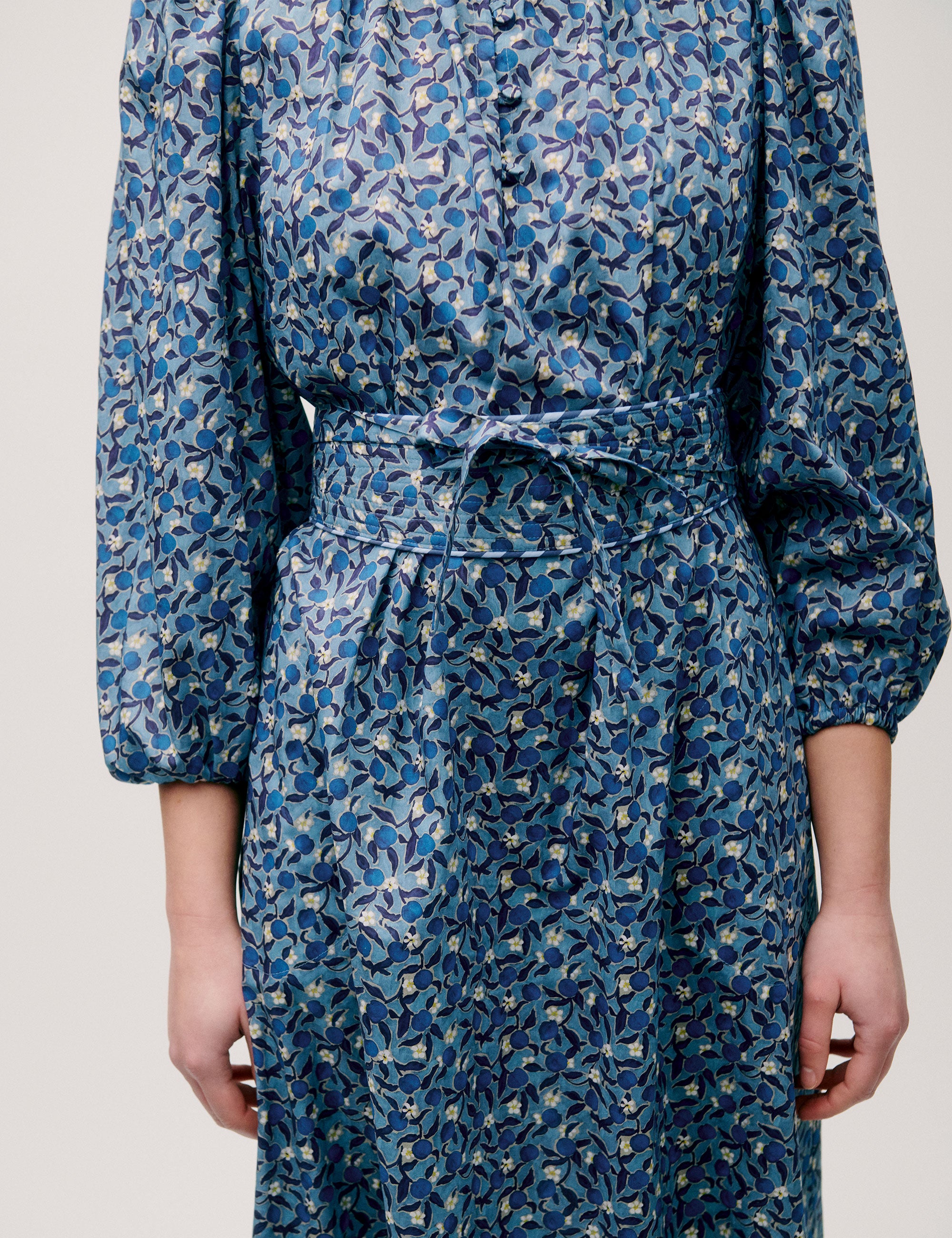 Woman wearing a long blue floral dress with puff sleeves, pleated neckline, and tie waist