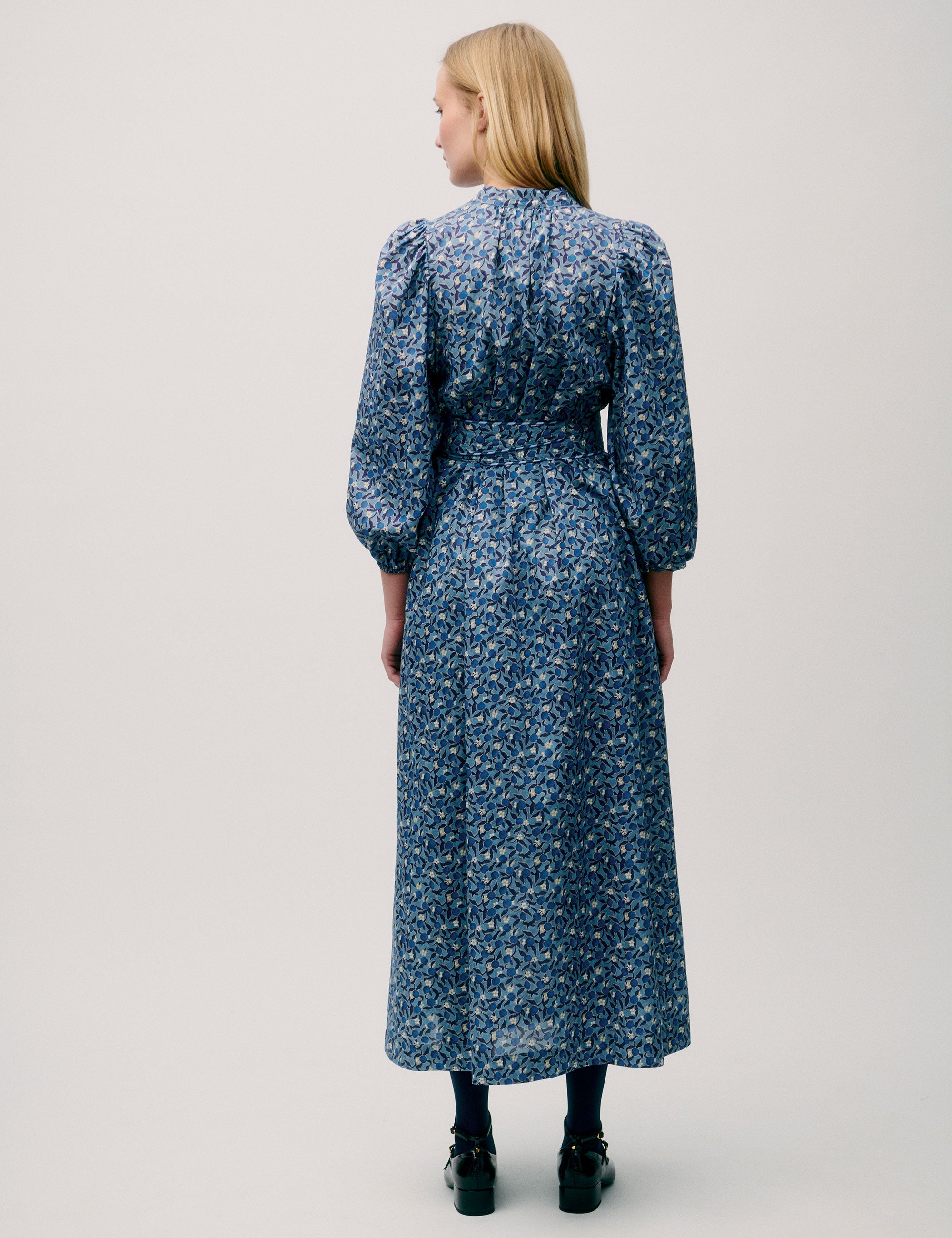 Woman wearing a long blue floral dress with puff sleeves, pleated neckline, and tie waist