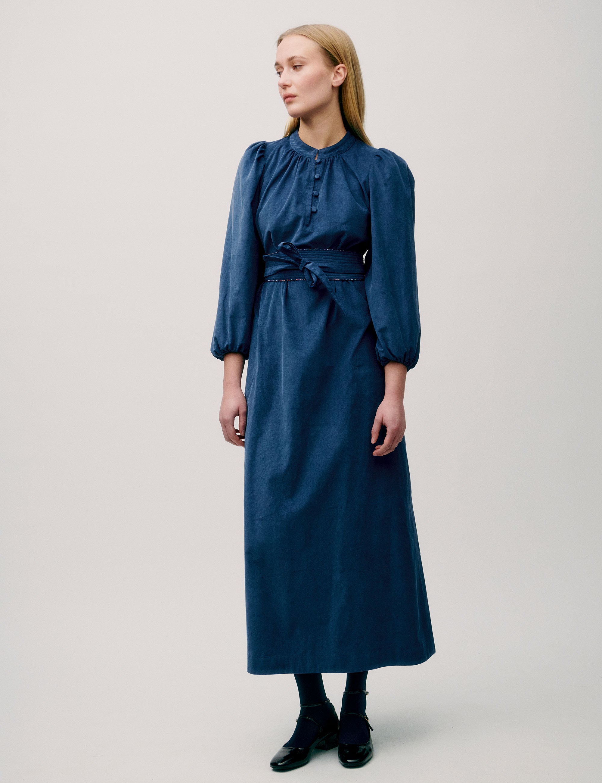 Woman wearing a long blue corduroy dress with puff sleeves, pleated neckline, and fabric waist tie