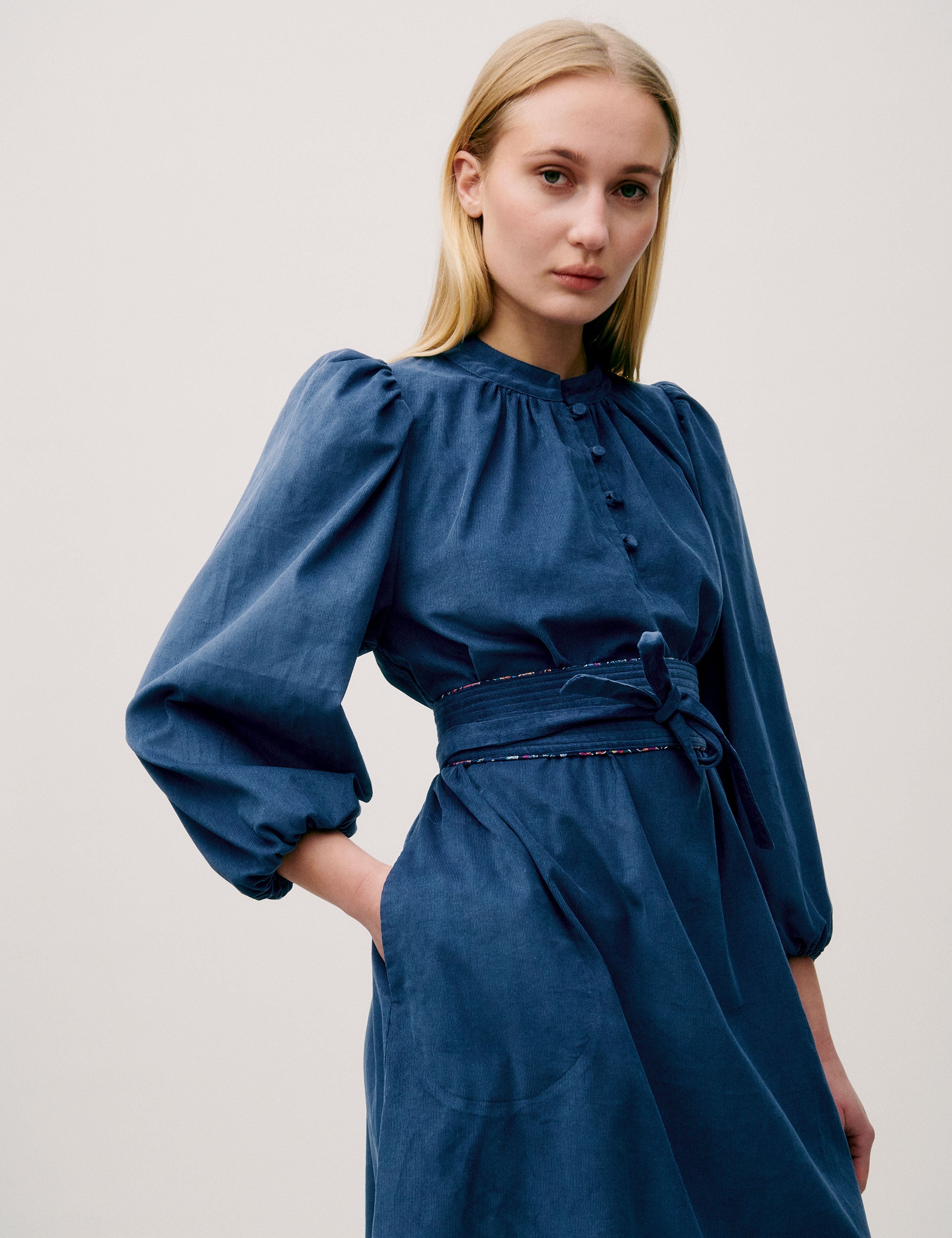 Woman wearing a long blue corduroy dress with puff sleeves, pleated neckline, and fabric waist tie