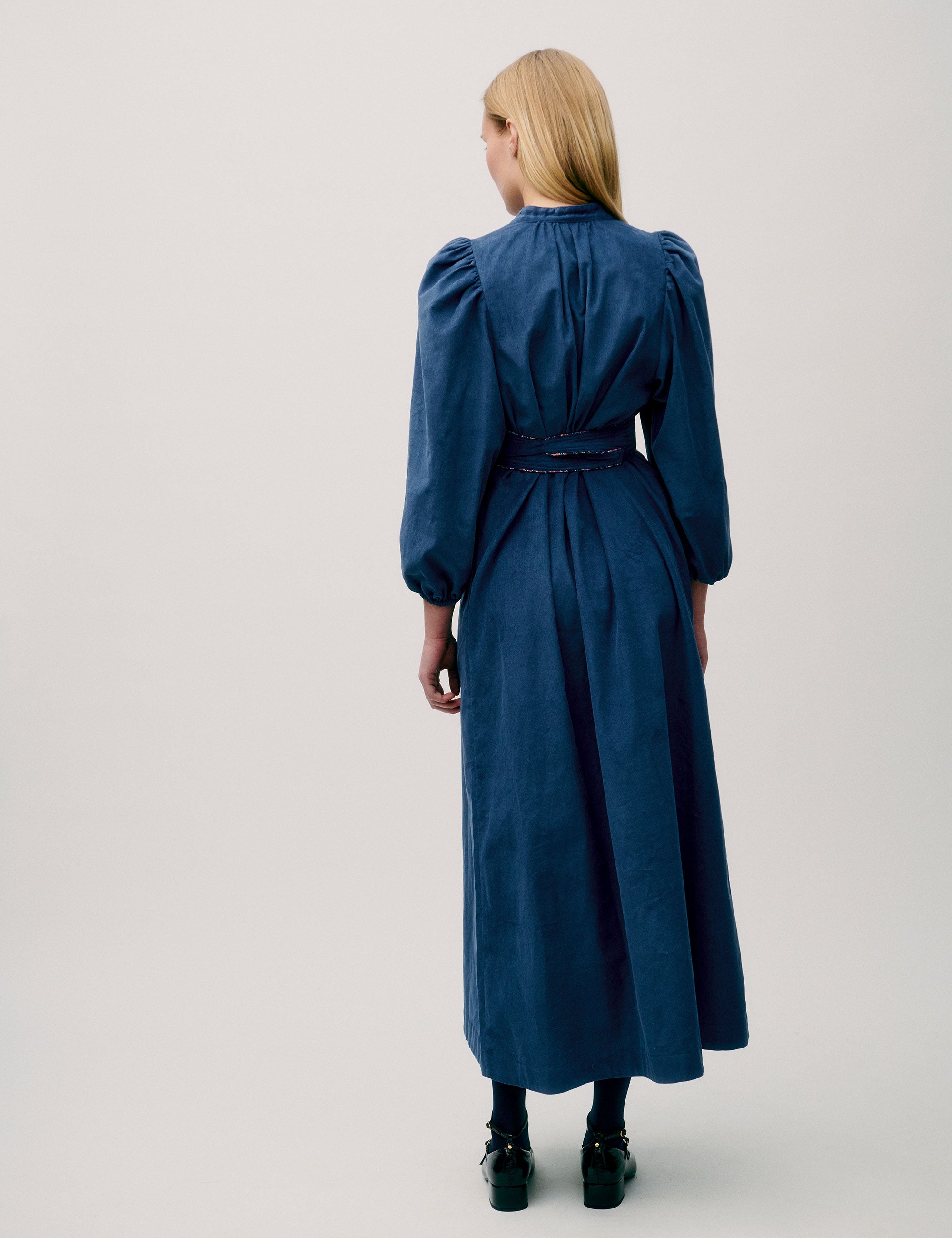 Woman wearing a long blue corduroy dress with puff sleeves, pleated neckline, and fabric waist tie